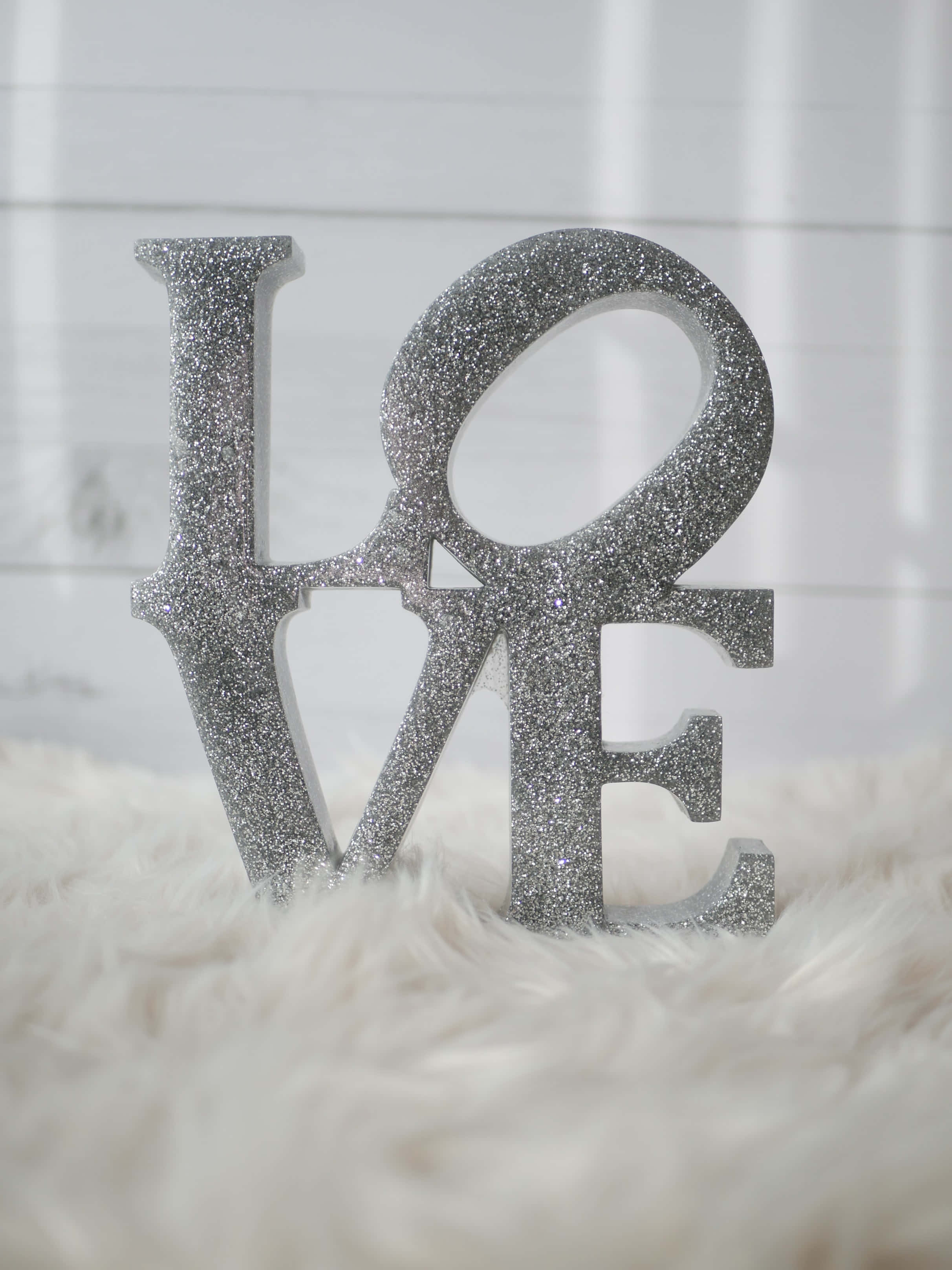 Silver Sparkle 桌布 壁紙 — 一個閃亮的銀色「LOVE」字雕塑優雅地立在白色毛絨地毯上，背景是明亮的白色護牆板牆壁。