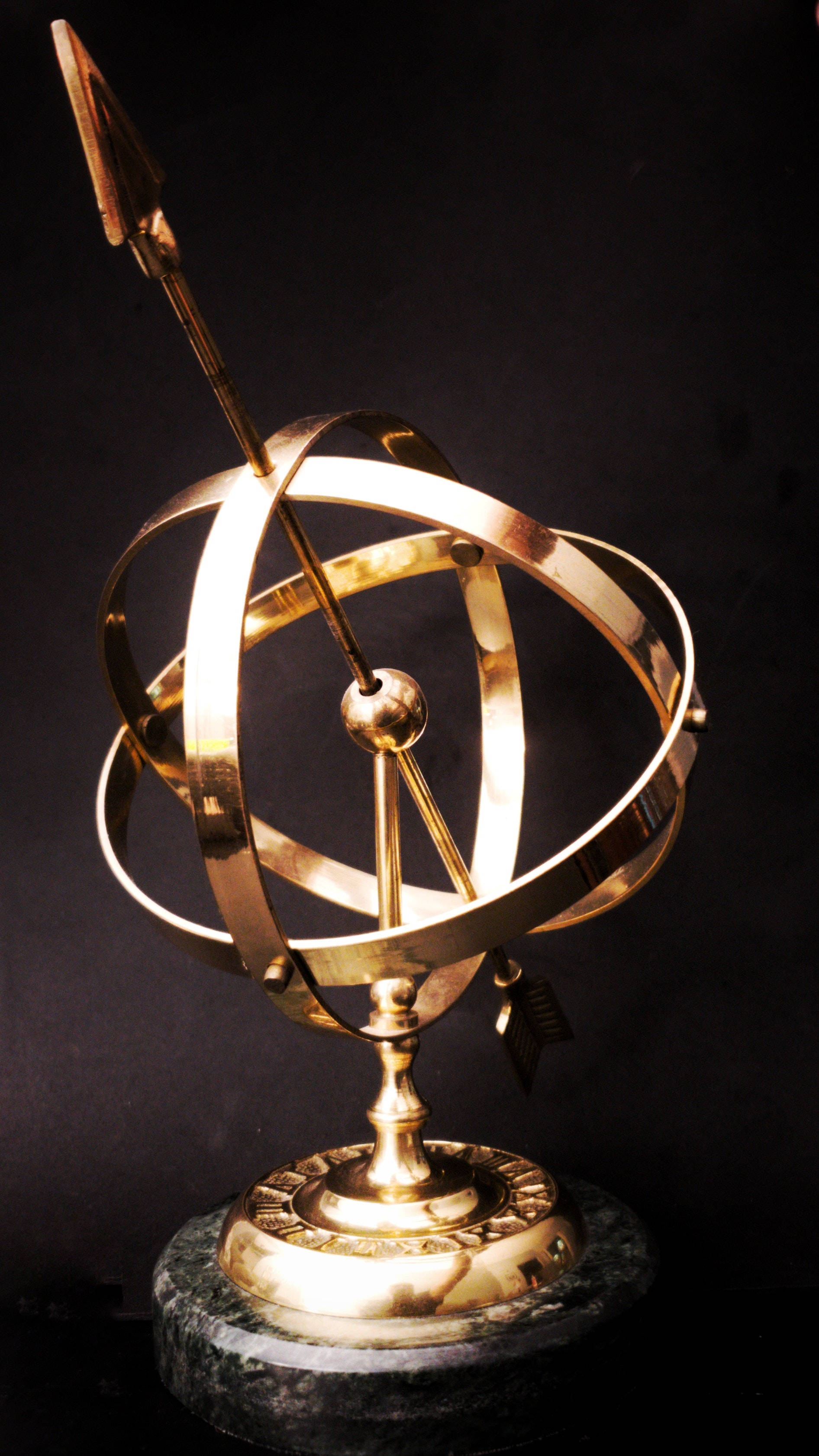 Gold Jewellery 桌布 壁紙 — 一個閃亮的黃銅渾天儀，帶有尖銳的日晷指針，立在黑色背景前的深色大理石底座上。