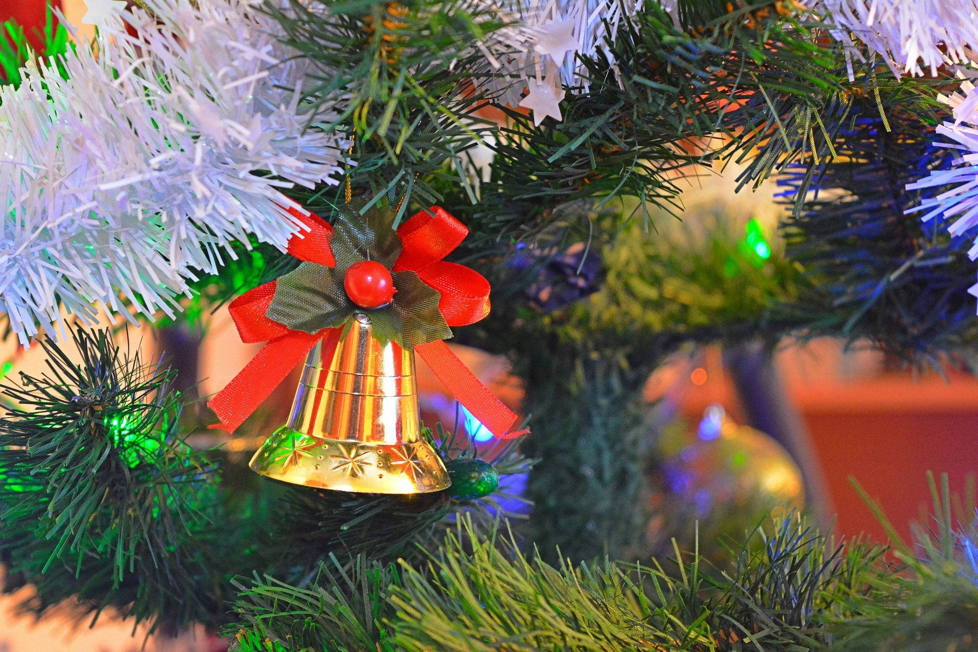 Christmas Bell 桌布 壁紙 — 一個閃亮金色鈴鐺裝飾品，帶有紅色蝴蝶結，掛在帶有發光燈飾的綠色和白色聖誕樹上的特寫。