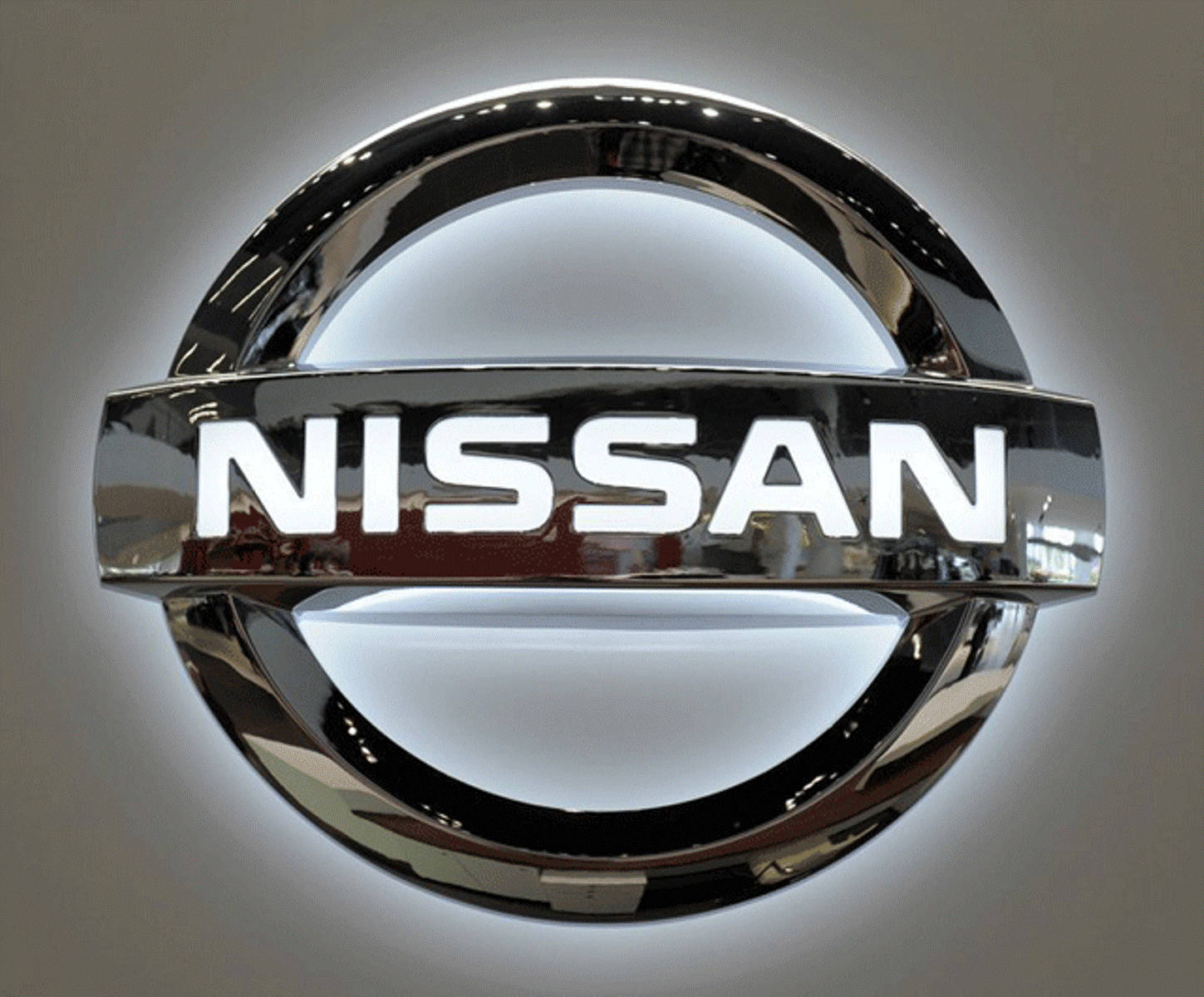 日產 桌布 壁紙 — 一個閃亮鍍鉻日產標誌的特寫，帶有發光的白色背光，背景是柔和的灰色牆壁。