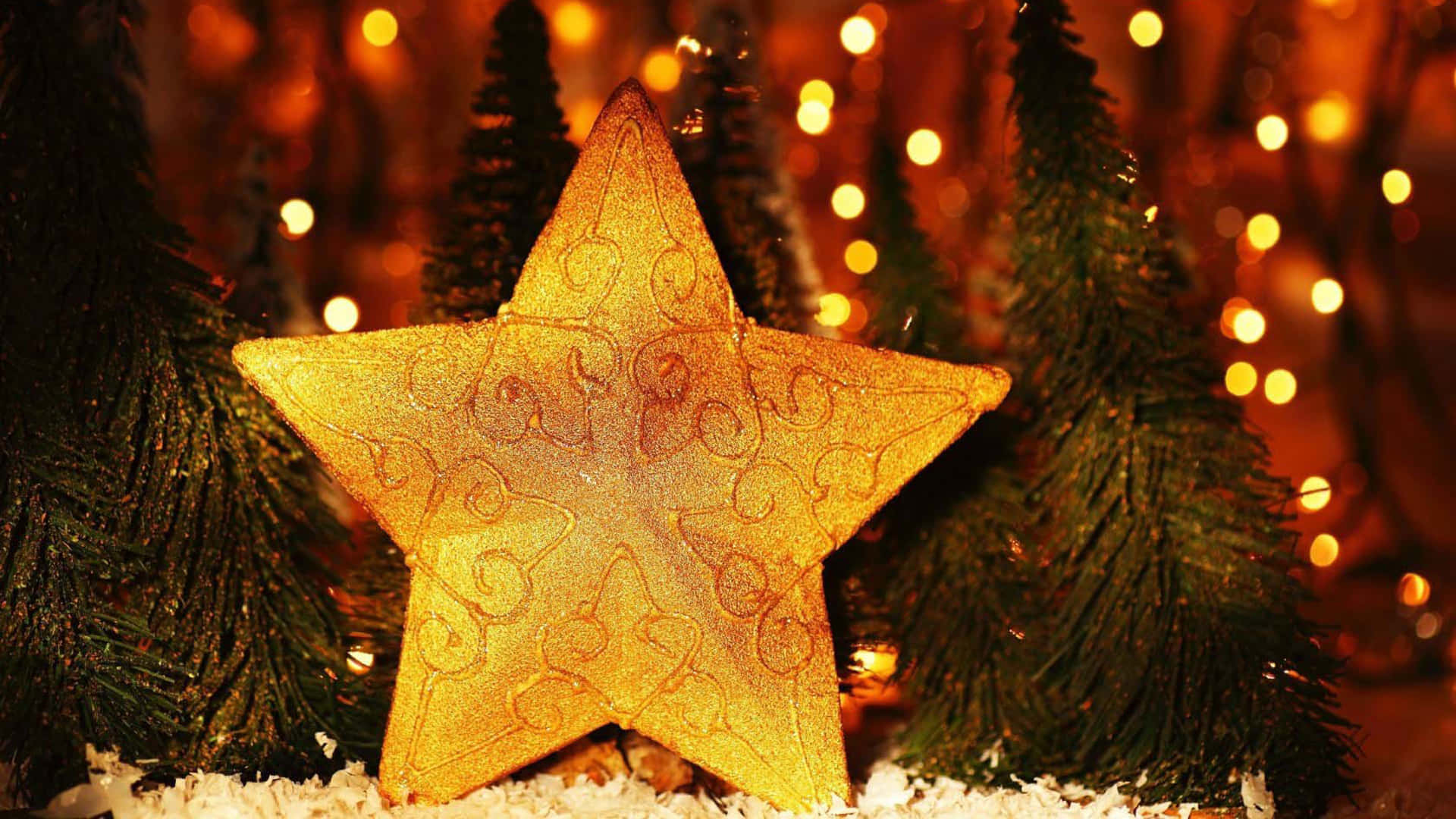 Christmas Star 桌布 壁紙 — 一個閃爍金色光芒、帶有複雜漩渦紋的星星裝飾品，放置在雪地上，周圍環繞著微型松樹和溫暖的散景燈光。