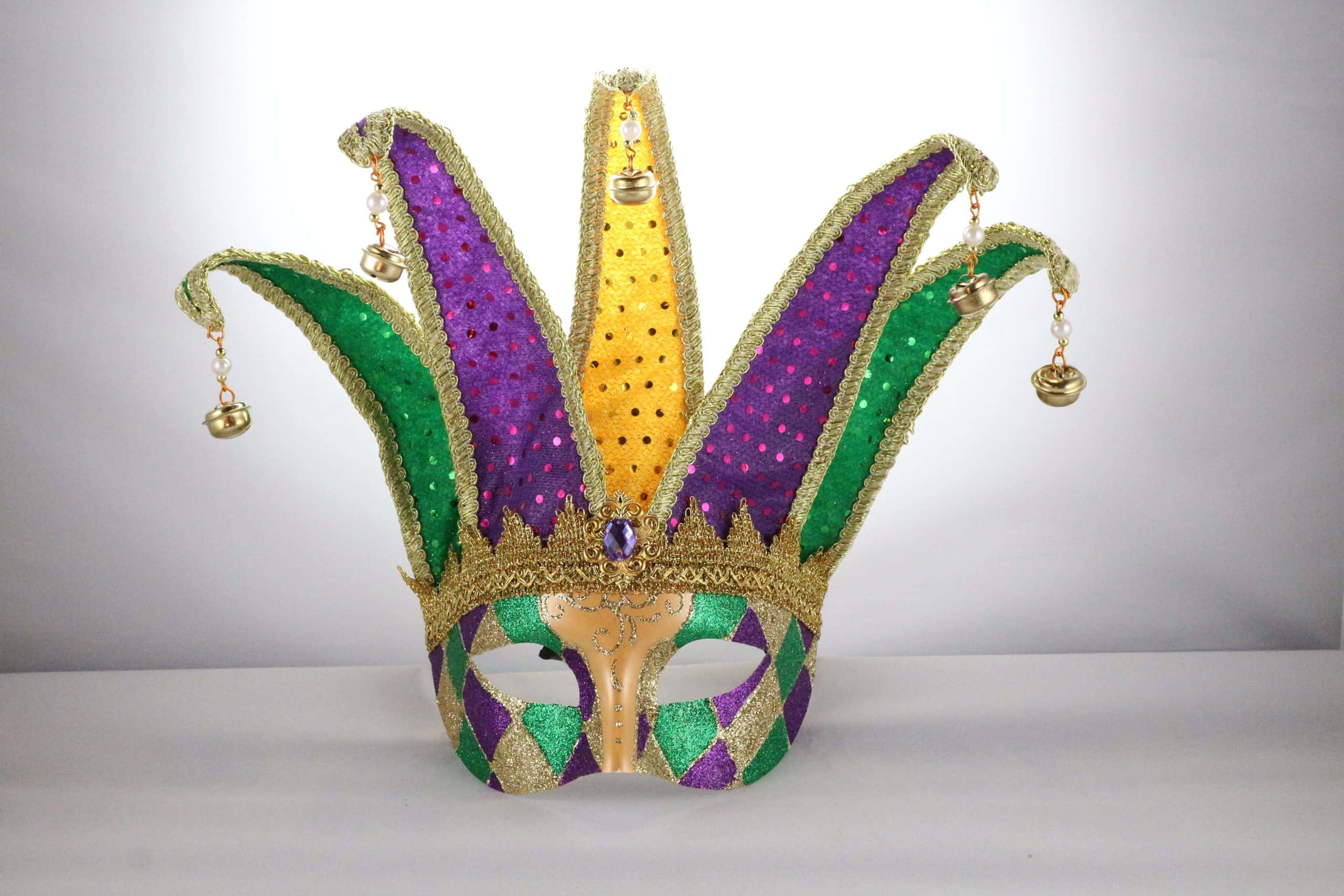 Mardi Gras 桌布 壁紙 — 一個閃閃發光的小丑面具，帶有紫色、綠色和金色的亮片以及懸掛的鈴鐺，放在白色表面上。