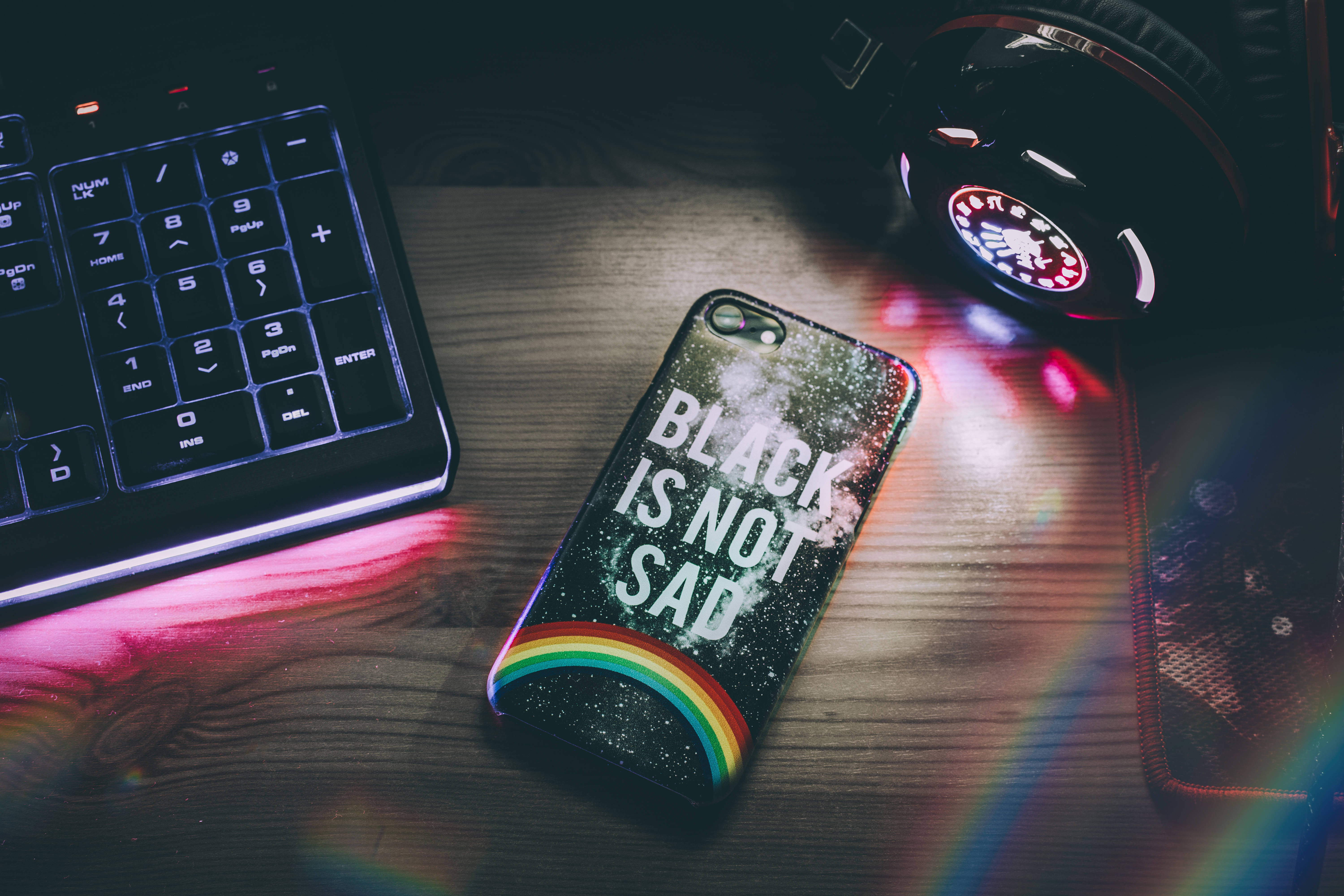 dark neon iPhone 桌布 壁紙 — 一個閃閃發光的手機殼，帶有彩虹和文字，放在木桌上，旁邊有鍵盤和耳機，在色彩繽紛的光暈下。