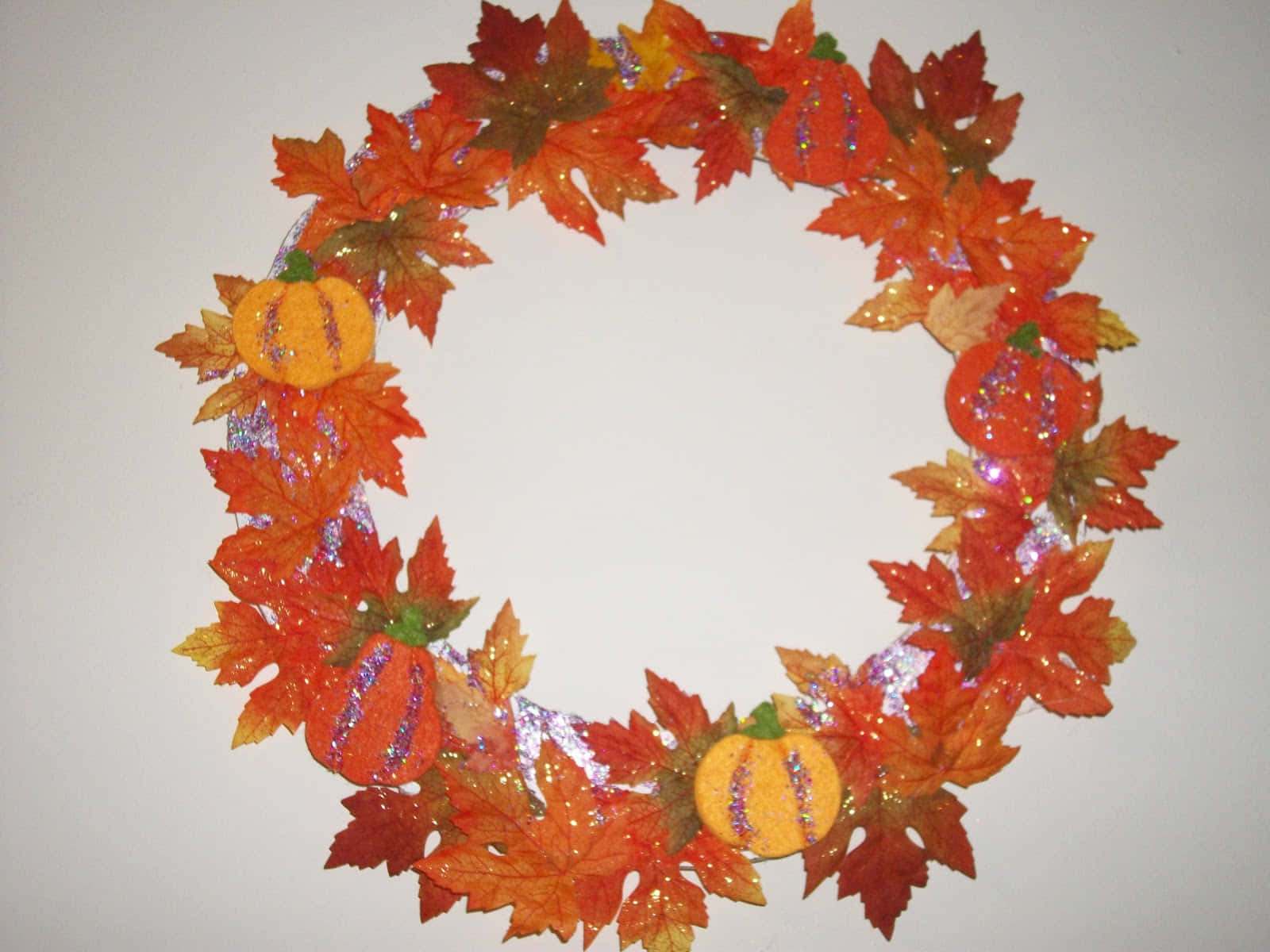 Fall Crafts 桌布 壁紙 — 一個閃閃發光的秋季花環，裝飾著橙色南瓜和鮮豔的楓葉，掛在素牆上。