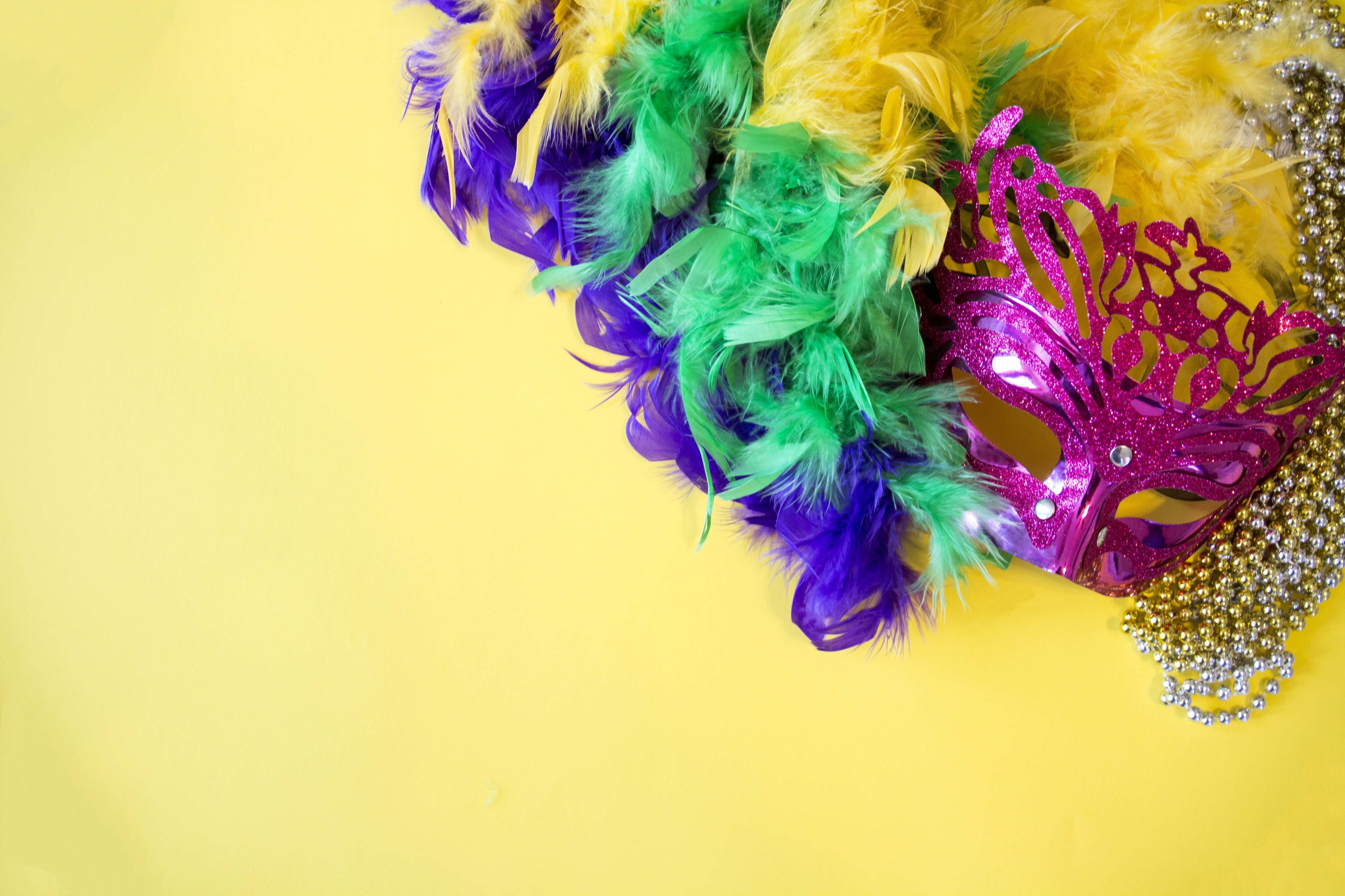 Mardi Gras 桌布 壁紙 — 一個閃閃發光的粉紅色狂歡節面具，鑲嵌在充滿活力的紫色、綠色和黃色羽毛以及金色珠子之間，背景為明亮的黃色。