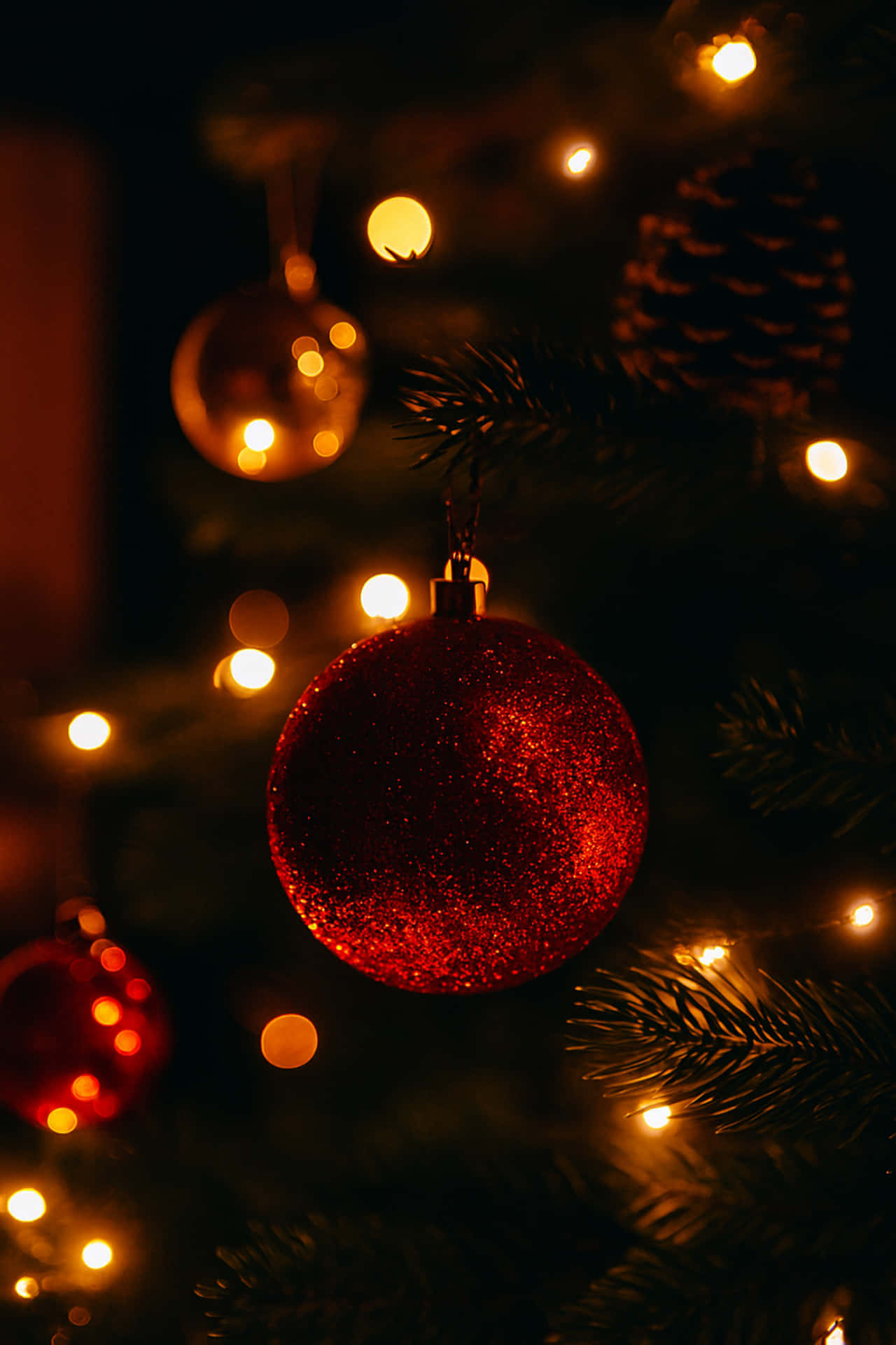 Christmas tree decorations 壁紙 — 一個閃閃發光的紅色裝飾品掛在黑暗的松樹枝上，被溫暖的金色仙女燈照亮。