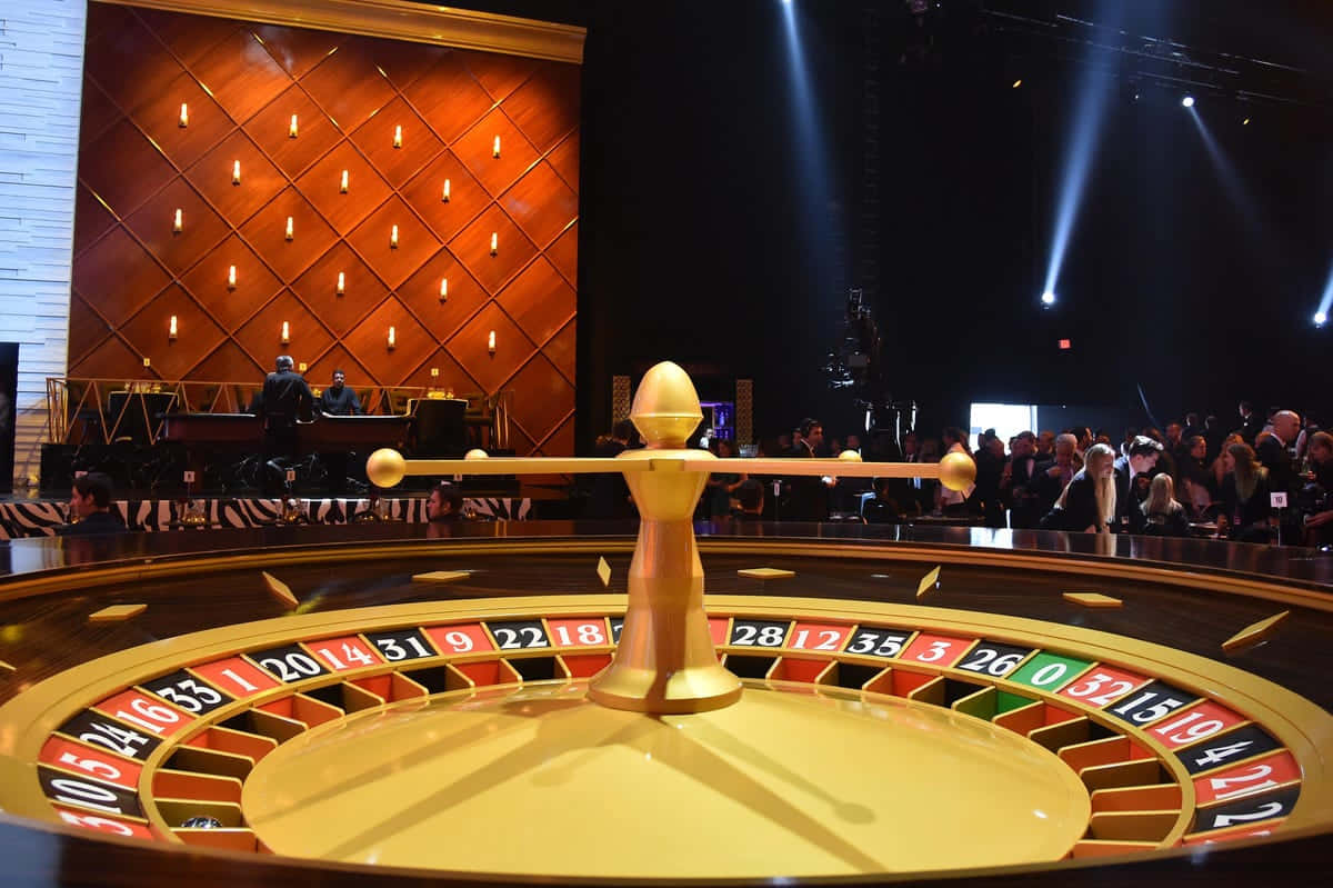 Casino 桌布 壁紙 — 一個閃閃發光的賭場輪盤在戲劇性的舞台燈光下佔據前景，觀眾專注地觀看。