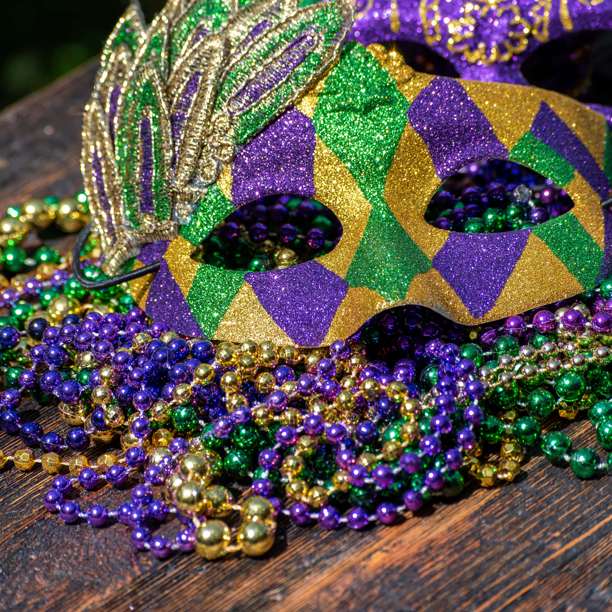 Mardi Gras 桌布 壁紙 — 一個閃閃發光的金色、綠色和紫色狂歡節面具放在一堆節日珠子上，置於風化的木質表面。