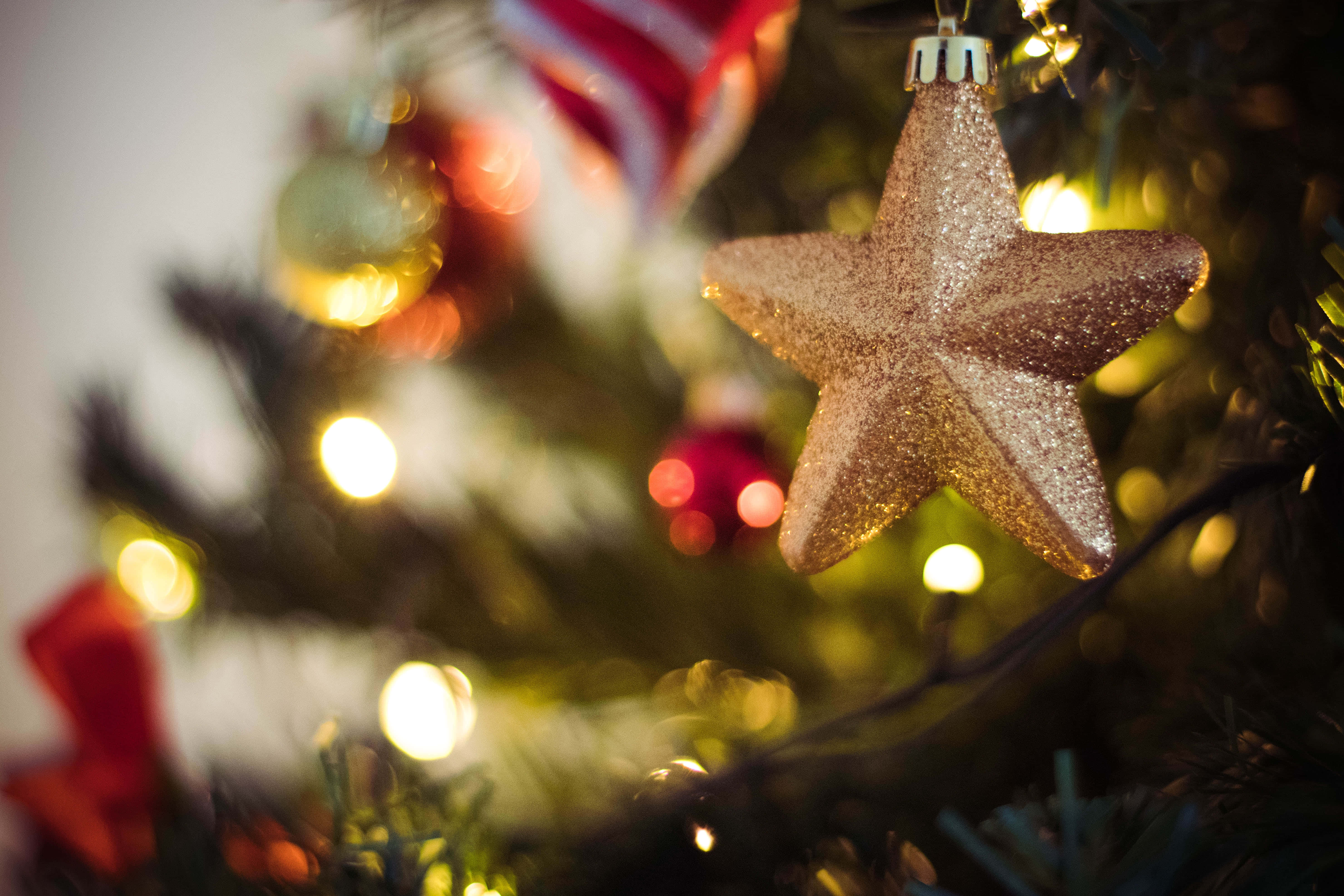 Christmas Star 桌布 壁紙 — 一個閃閃發光的金色星星裝飾品在焦點中懸掛，背景是柔和模糊的聖誕樹，帶有溫暖的燈光和紅色點綴。