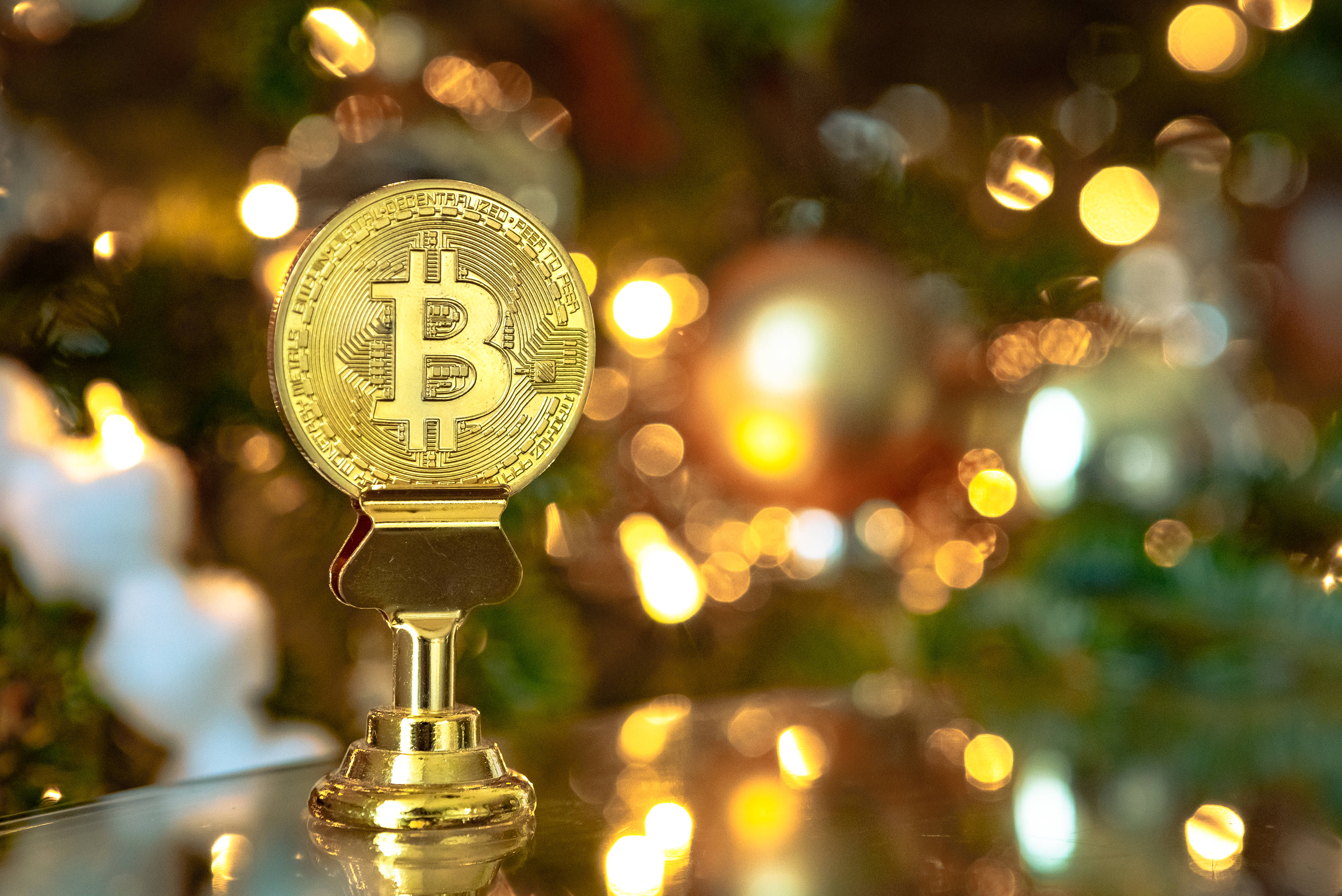 比特幣 桌布 壁紙 — 一個閃閃發光的金色比特幣獎盃立於反光表面上，背景是節日燈飾和綠化的散景效果。