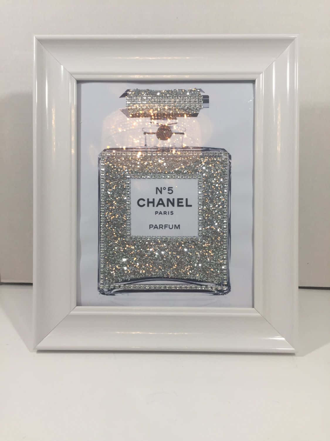 Chanel 桌布 壁紙 — 一個閃閃發光的銀色Chanel No 5香水瓶藝術品，白色框架，展示在素牆上。