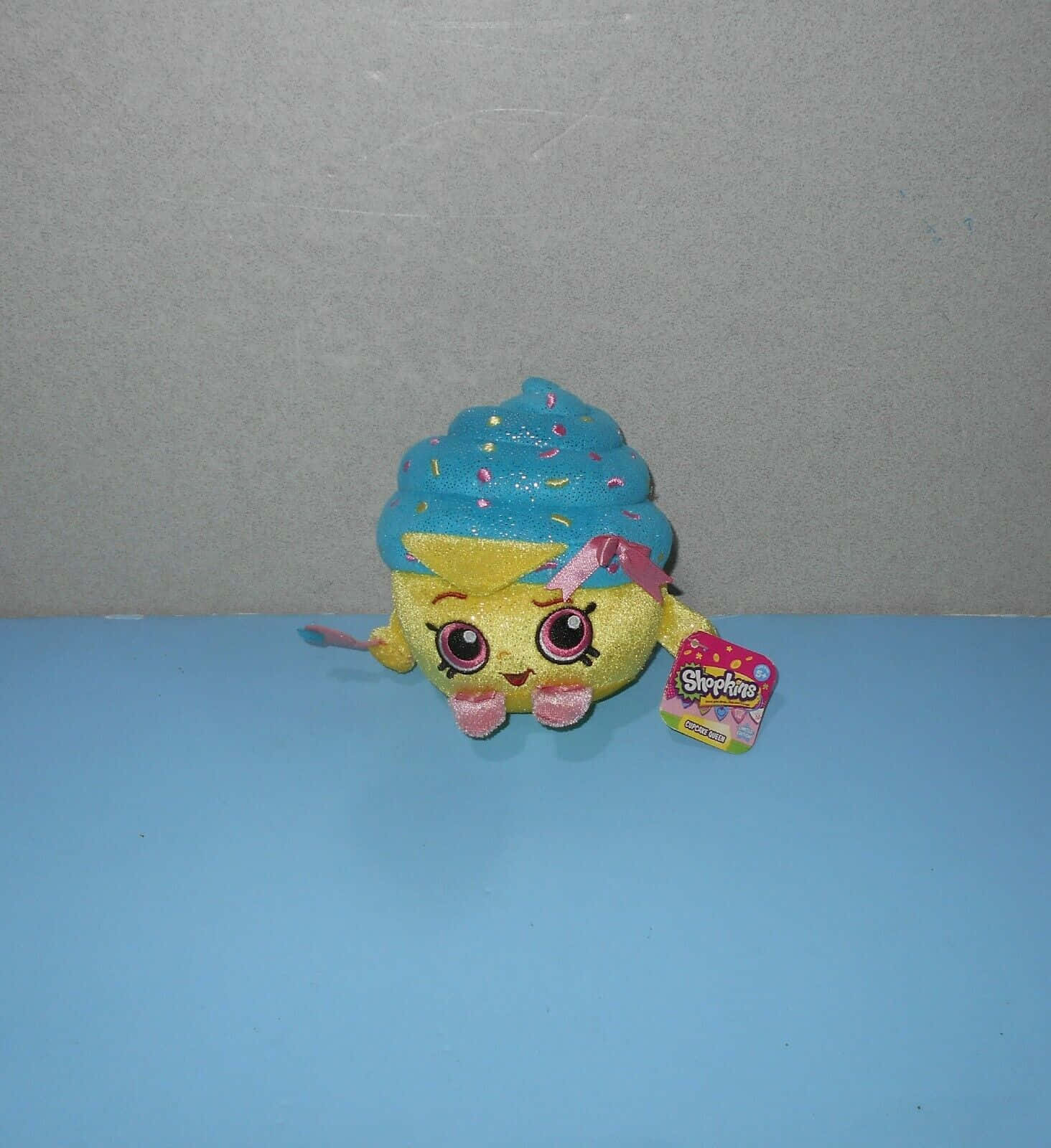Shopkins 桌布 壁紙 — 一個閃閃發光的黃色杯子蛋糕絨毛玩具，帶有藍色糖霜和粉紅色糖粒，放在藍色表面上，背景是灰色牆壁。
