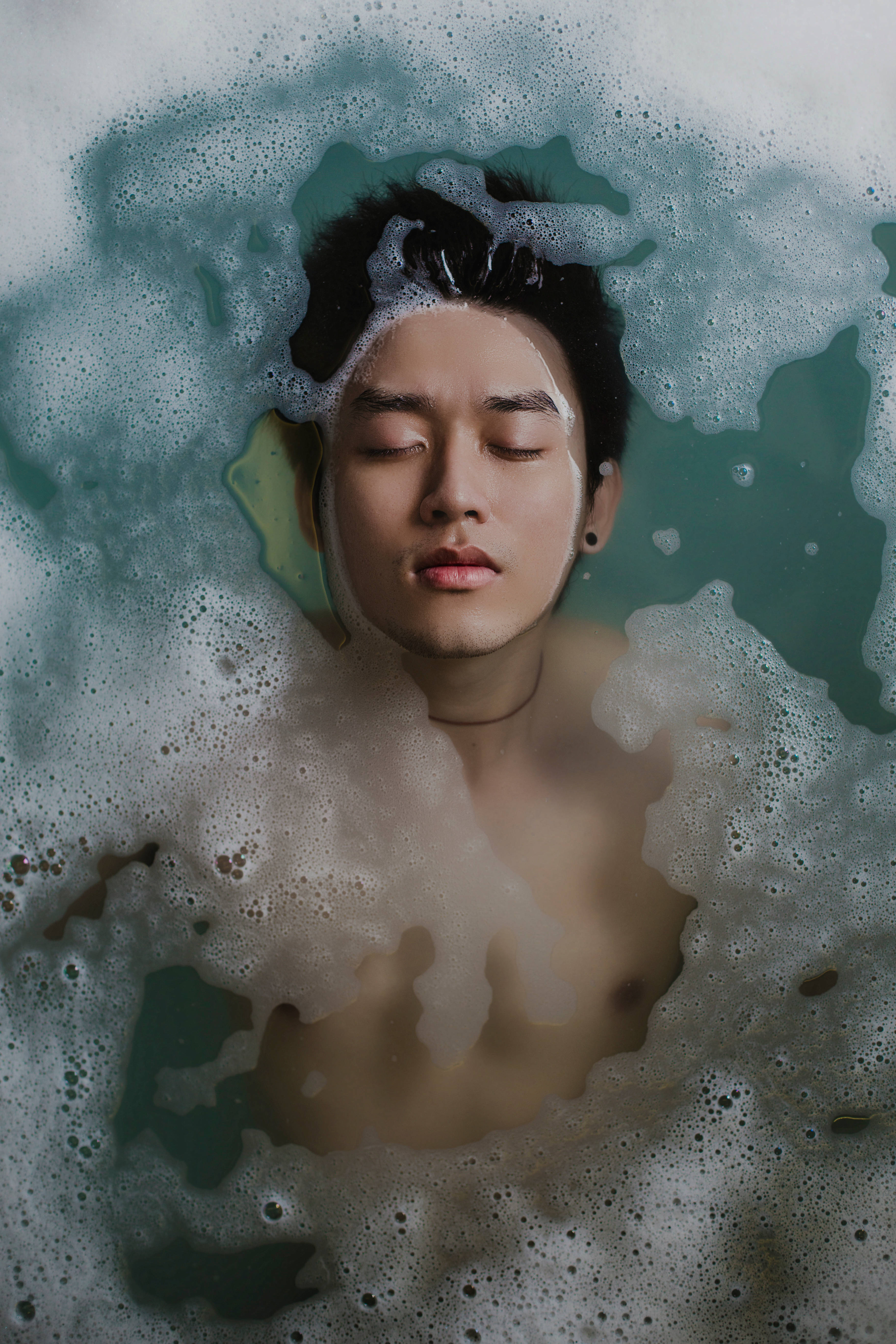Sexy 桌布 壁紙 — 一個閉上眼睛的年輕男子平靜地漂浮在一個充滿旋轉藍綠色和白色泡泡的浴缸中。