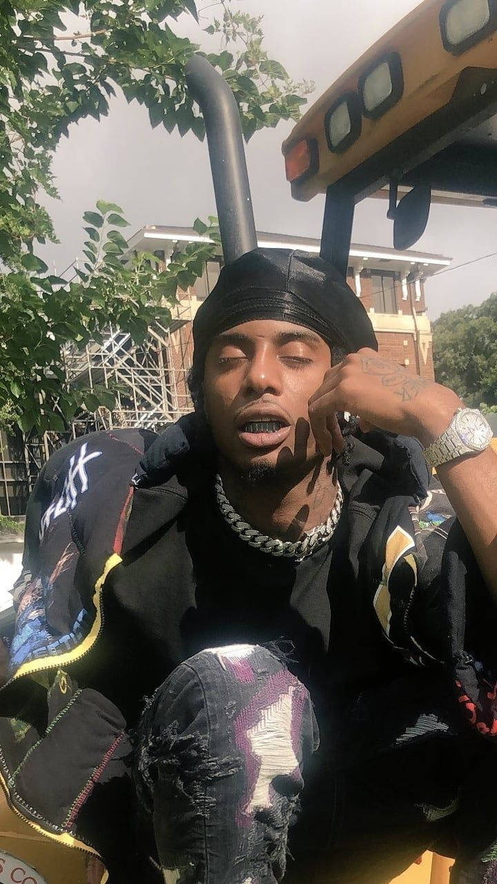 Carti 桌布 壁紙 — 一個閉著眼睛、戴著銀鏈的年輕男子坐在黃色工程車裡，車停在枝葉茂密的樹下。