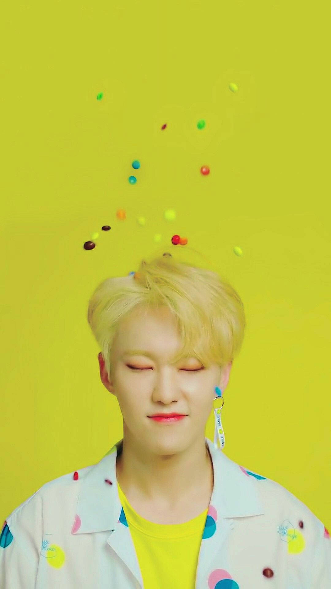 Hoshi 桌布 壁紙 — 一個閉著眼睛、金髮的年輕男子在明亮的黃色背景前微笑，彩色糖果漂浮在他的頭周圍。