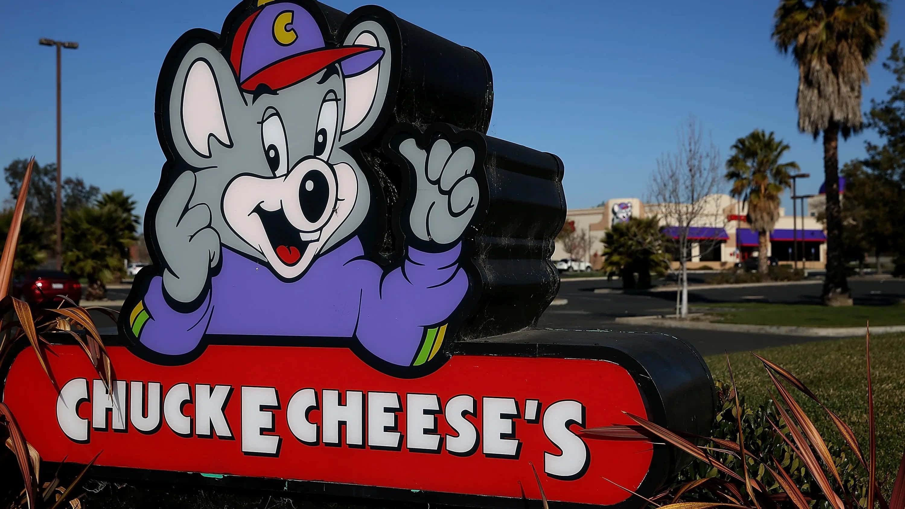 查克芝士 桌布 壁紙 — 一個開心的 Chuck E Cheese 吉祥物雕像在藍天白雲、棕櫚樹和餐廳背景下，從紅色招牌上揮手。