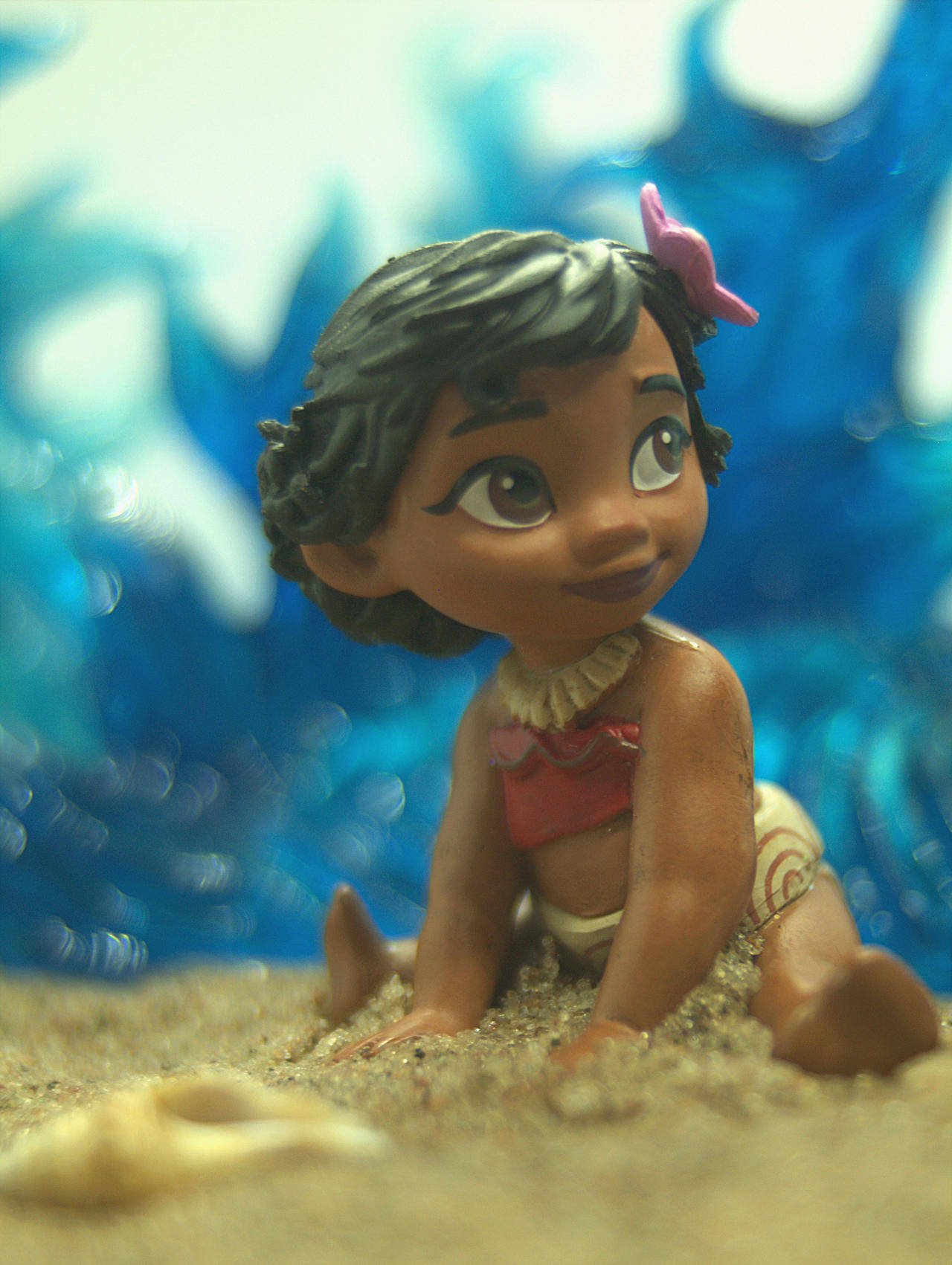 Baby Moana 桌布 壁紙 — 一個開心的Moana公仔坐在沙灘上，背後是模糊的藍色海浪。