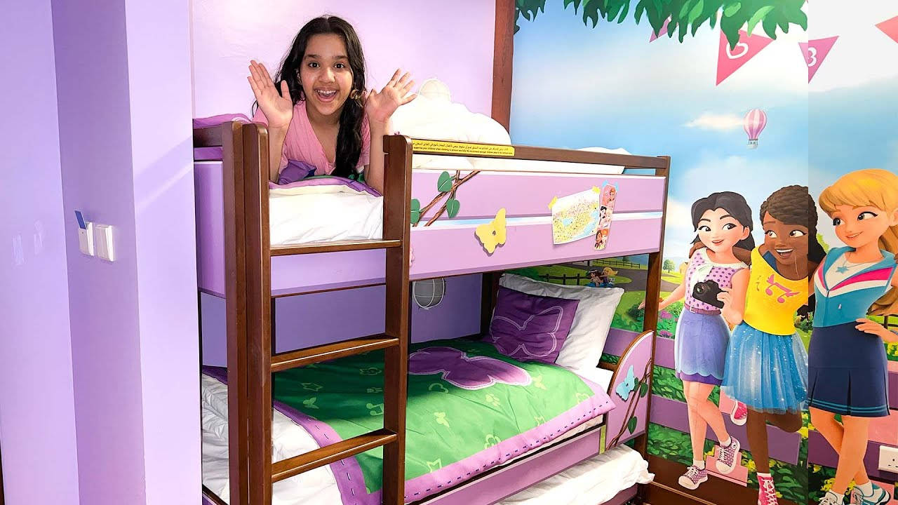 Bedroom 桌布 壁紙 — 一個開心的女孩在紫色雙層床上揮手，房間是充滿活力的卡通主題兒童房，有色彩繽紛的牆貼。