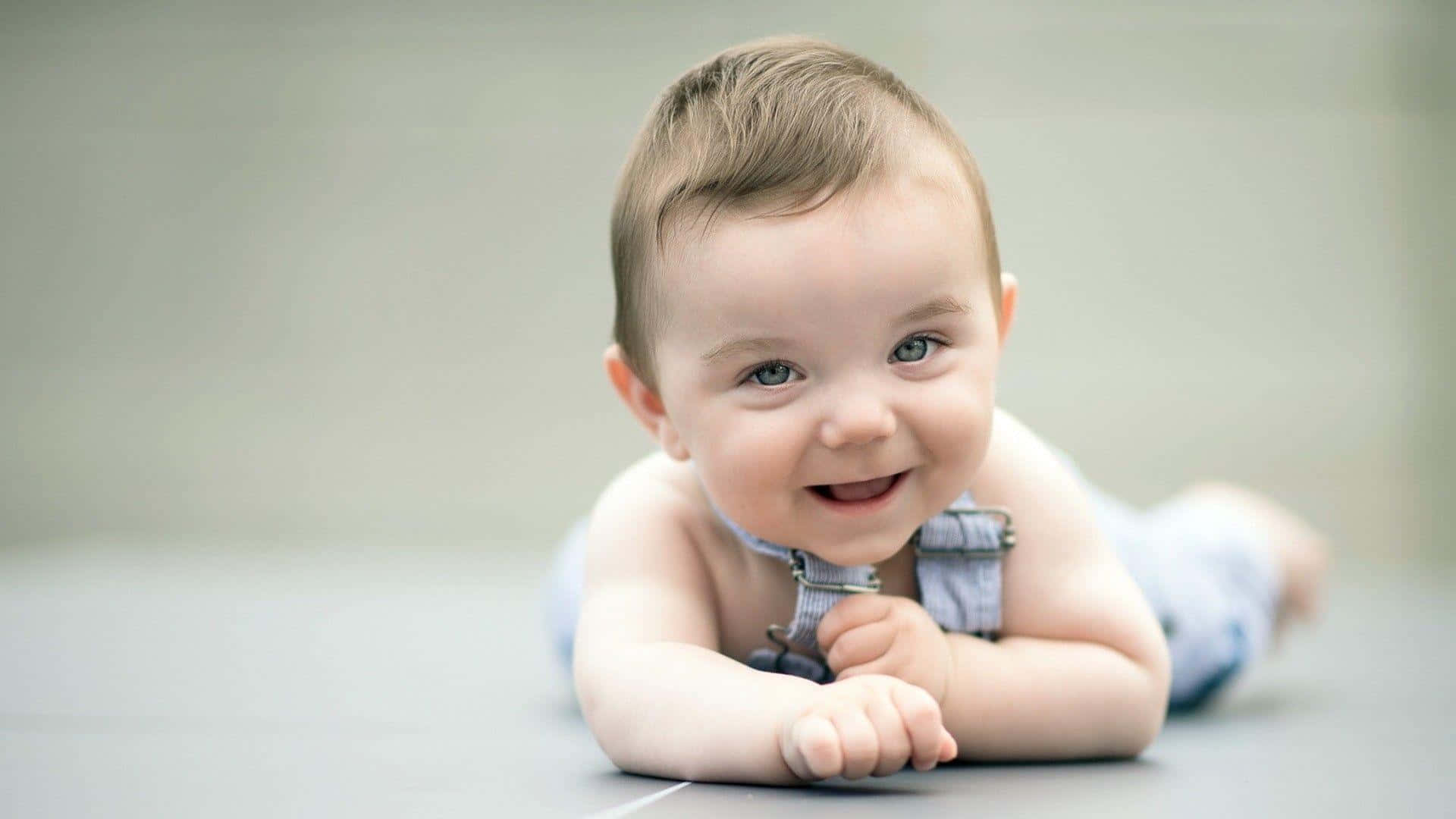 cute baby 桌布 壁紙 — 一個開心的嬰兒，有著明亮的藍眼睛，趴著躺在光滑的灰色地板上微笑。