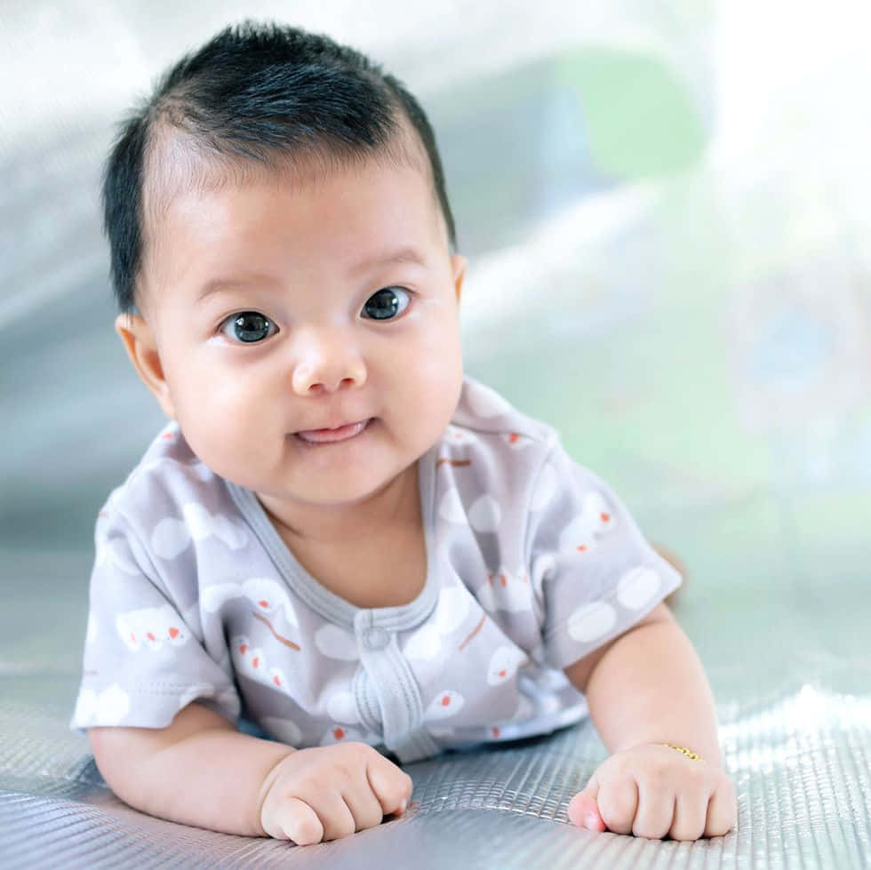 Baby Boy 桌布 壁紙 — 一個開心的嬰兒，深色眼睛和短黑髮，躺在有紋理的銀色表面上微笑。