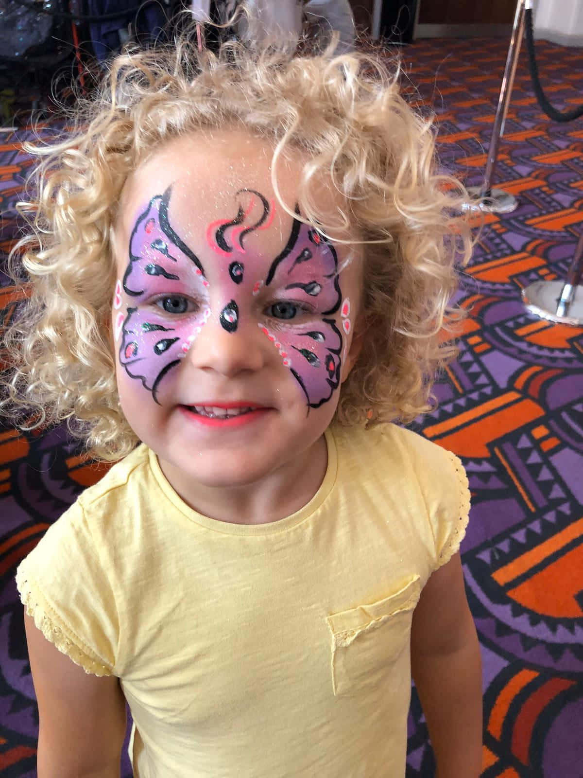 Butterfly Face Painting 桌布 壁紙 — 一個開心的孩子，有著捲曲的金色頭髮，在色彩繽紛的圖案地毯上，臉上畫著鮮豔的蝴蝶彩繪，笑容燦爛。