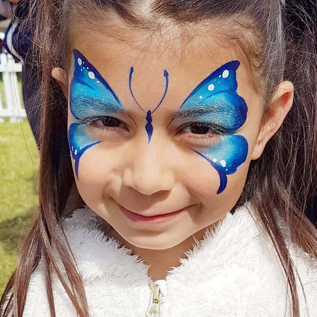 Butterfly Face Painting 桌布 壁紙 — 一個開心的孩子在陽光明媚的戶外背景下，帶著鮮豔的藍色蝴蝶臉部彩繪微笑。