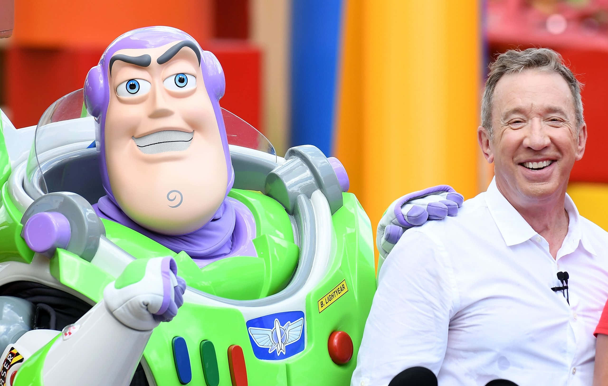 Buzz Lightyear 桌布 壁紙 — 一個開心的男人在色彩繽紛的環境中，微笑地站在一個大型巴斯光年玩具公仔旁邊。