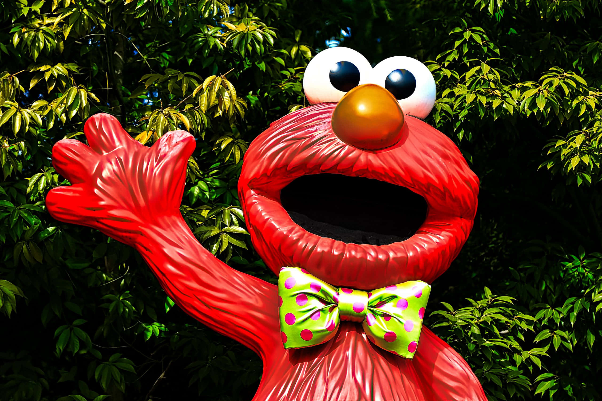 Elmo 桌布 壁紙 — 一個開心的紅色Elmo雕像，在茂密綠色樹葉背景下揮動一隻手。