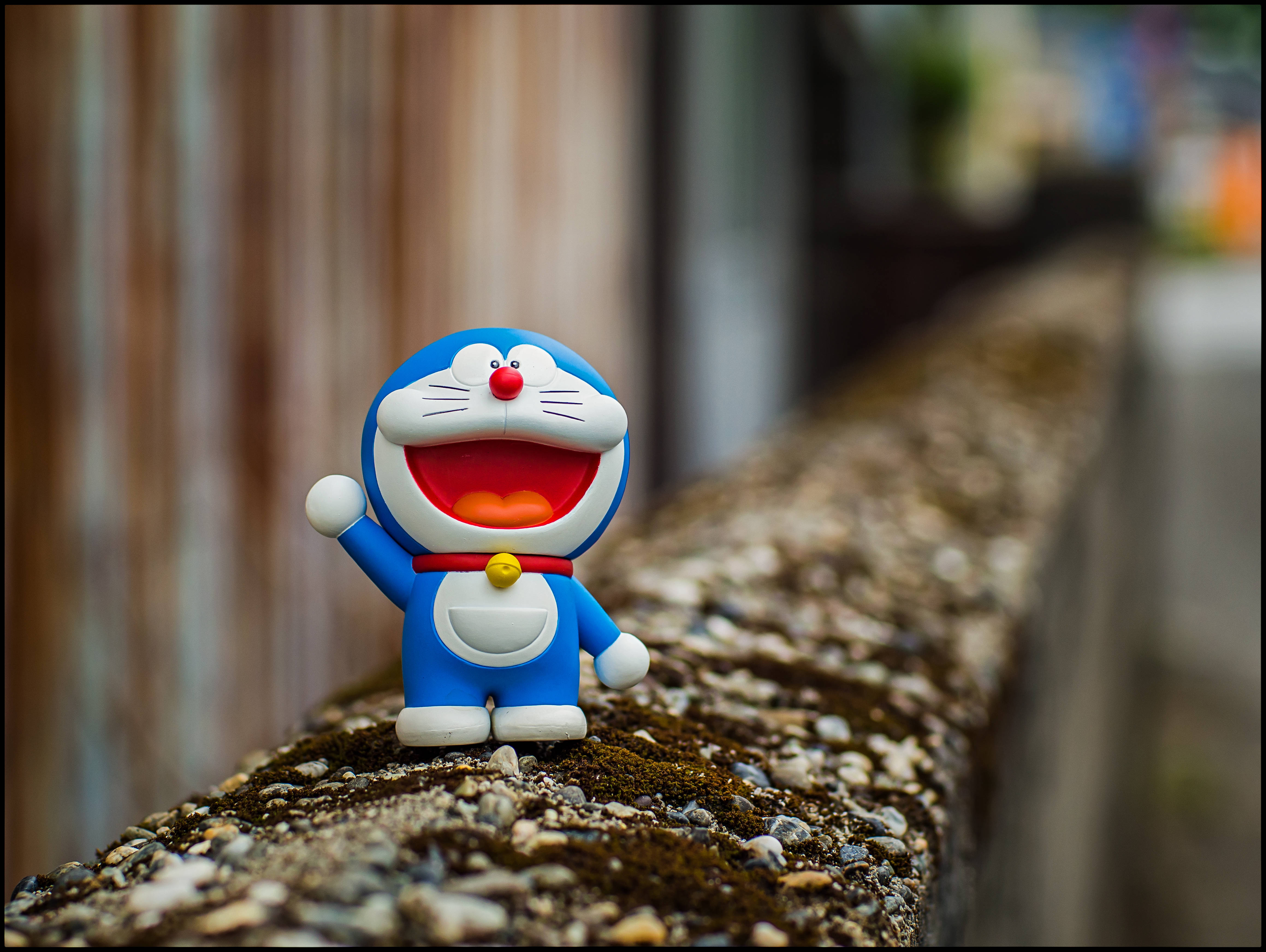 Doraemon 桌布 壁紙 — 一個開心的藍色哆啦A夢公仔，張大嘴巴，舉起一隻手臂，站在一個長滿苔蘚的石檯上，背景是模糊的木紋。
