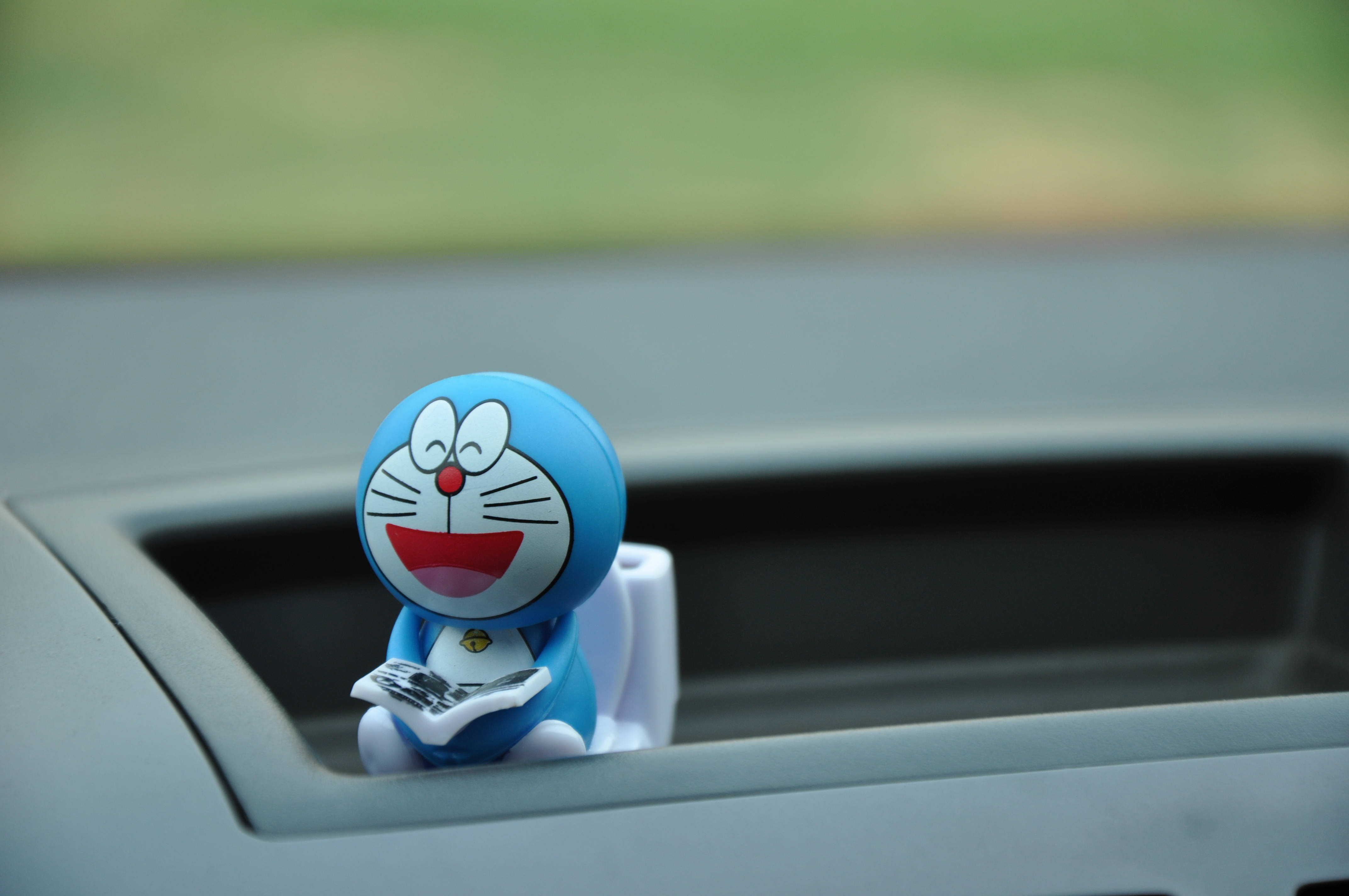 Doraemon 桌布 壁紙 — 一個開心的藍色哆啦A夢公仔坐在汽車杯架上閱讀報紙，背景是模糊的綠色。