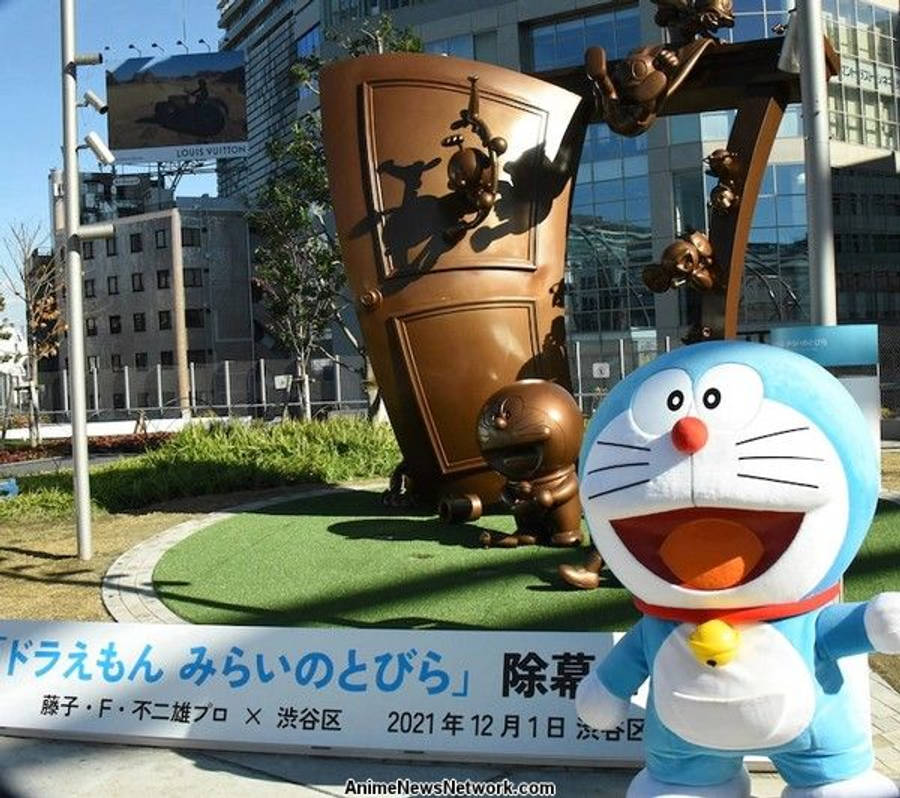 doraemon 4K 桌布 壁紙 — 一個開心的藍色機器人吉祥物站在一個陽光普照的城市廣場上，面前是一個帶有頑皮猴子圖案的銅門雕像。