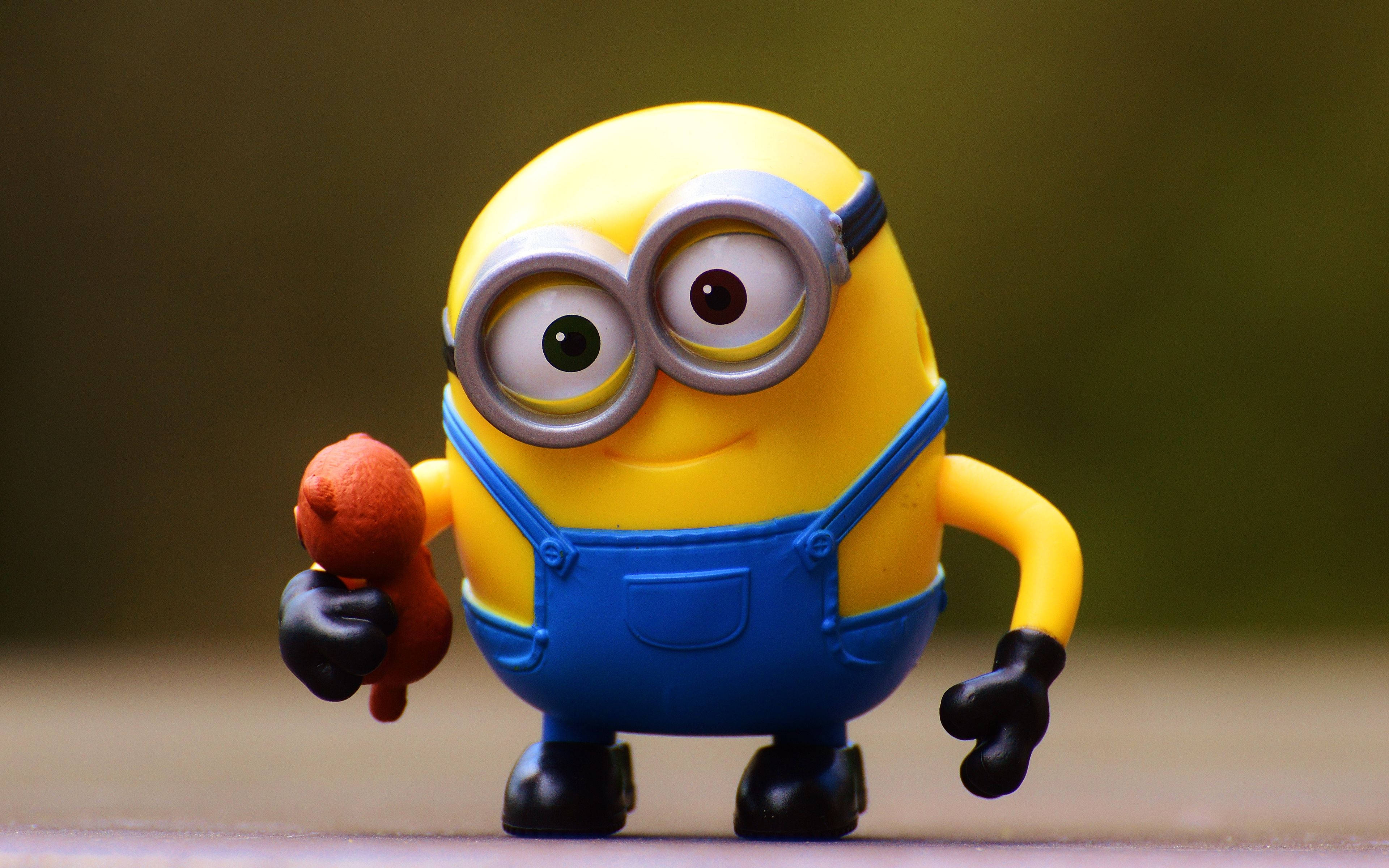Minion 桌布 壁紙 — 一個開心的黃色小小兵玩具，身穿藍色吊帶褲，在模糊的綠色背景前拿著一條香腸。