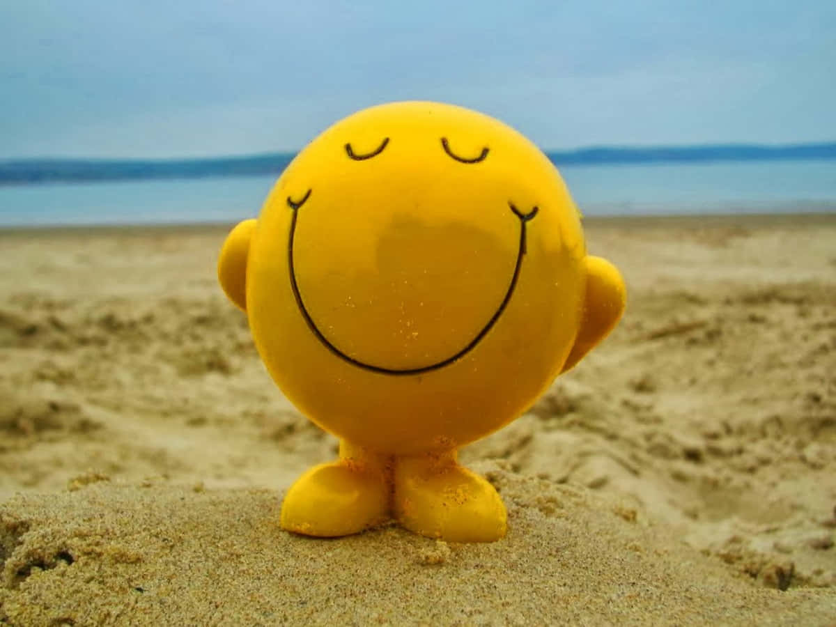Smiley 桌布 壁紙 — 一個開心的黃色笑臉公仔站在沙灘上，背景是平靜的海洋和多雲的天空。