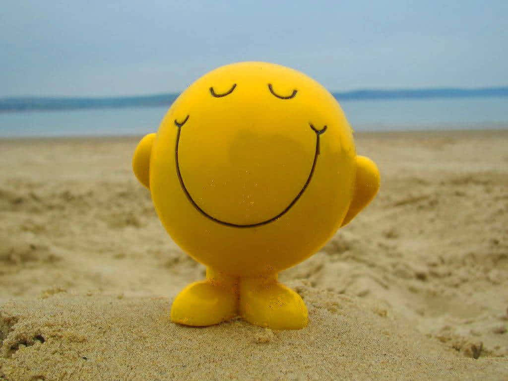 Happy Desktop 桌布 壁紙 — 一個開心的黃色笑臉玩具愉快地站在沙灘上，背景有平靜的水面和遙遠的地平線。