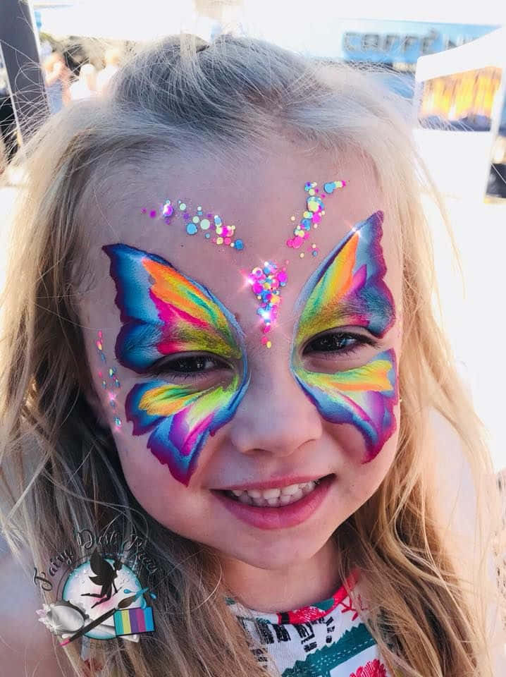 Butterfly Face Painting 桌布 壁紙 — 一個開朗的小女孩，臉上畫著鮮豔的彩虹蝴蝶彩繪和亮粉，對著鏡頭燦爛地微笑。