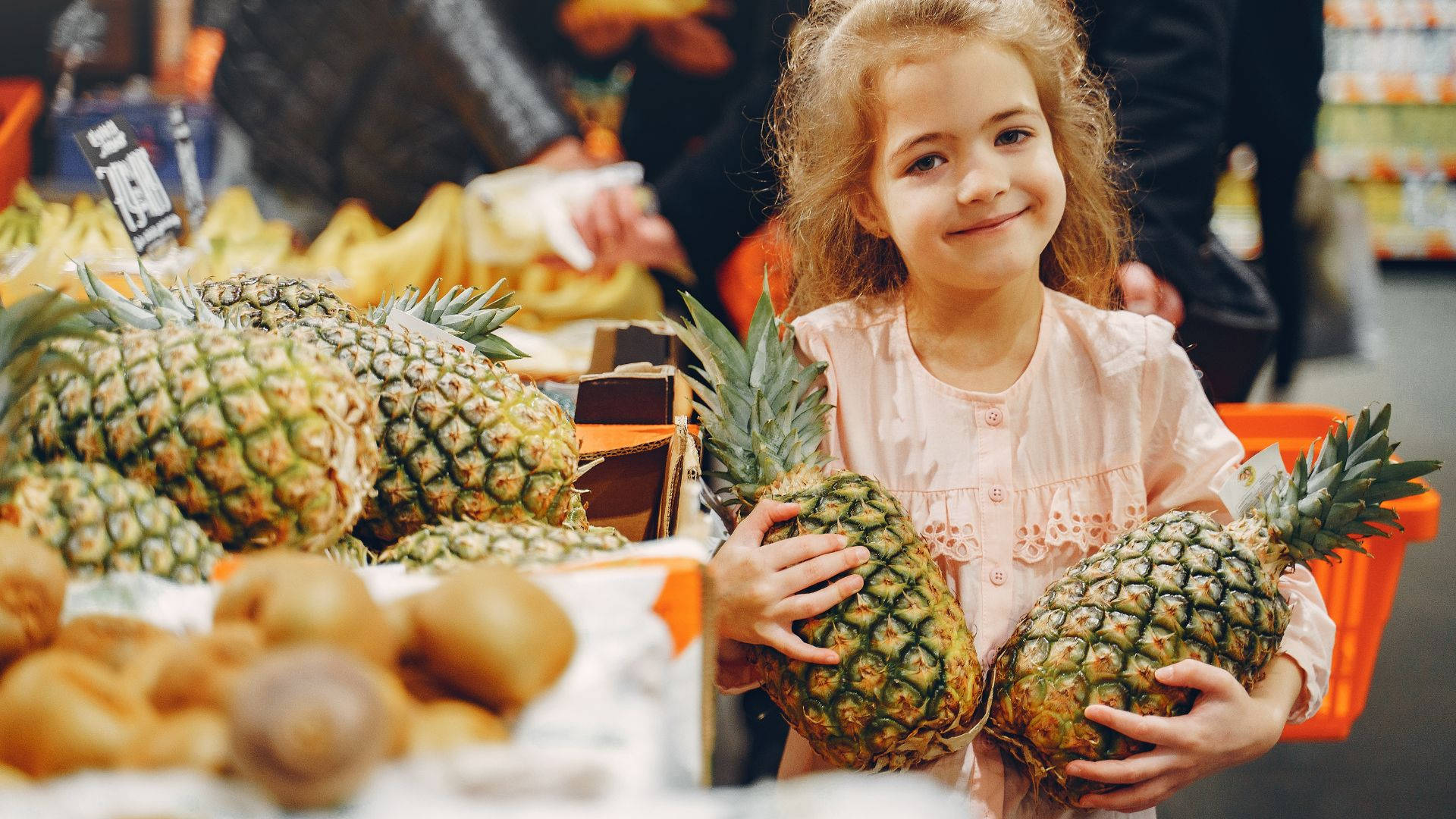 Pineapple 桌布 壁紙 — 一個開朗的小女孩在充滿活力的雜貨店農產品區拿著兩個鳳梨。