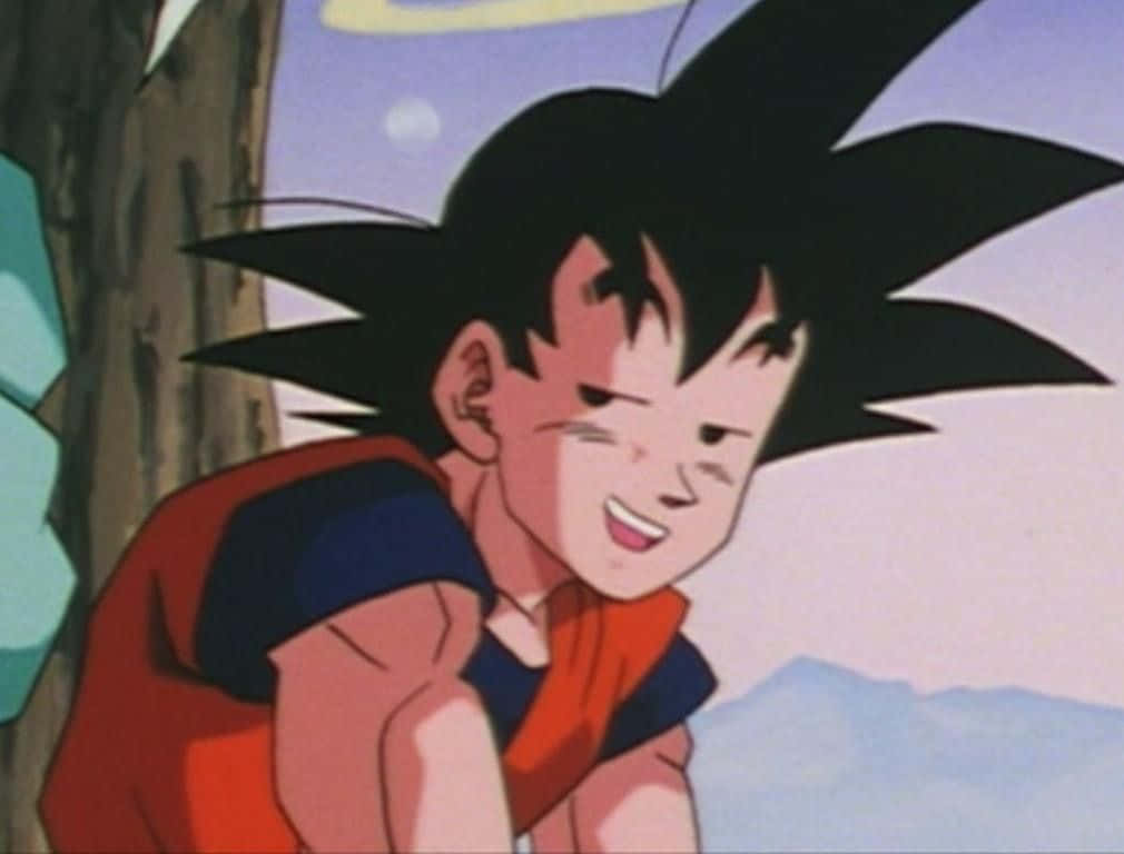 Funny Goku 桌布 壁紙 — 一個開朗的年輕動漫男孩，黑色刺蝟頭，穿著橙色道服，微笑著靠在山景中的一棵樹上。