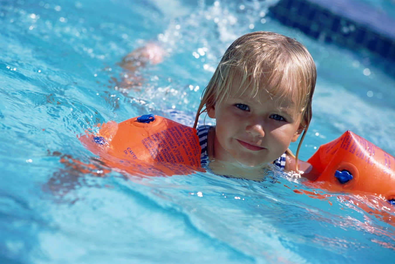 Swimming 桌布 壁紙 — 一個開朗的金髮小孩在閃閃發光的藍色游泳池中戴著亮橙色臂圈微笑。