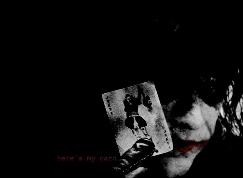 Joker Card 桌布 壁紙 — 一個陰暗的人物，紅唇，在黑色背景上舉起一張小丑撲克牌，紅色文字寫著「這是我的卡片」。