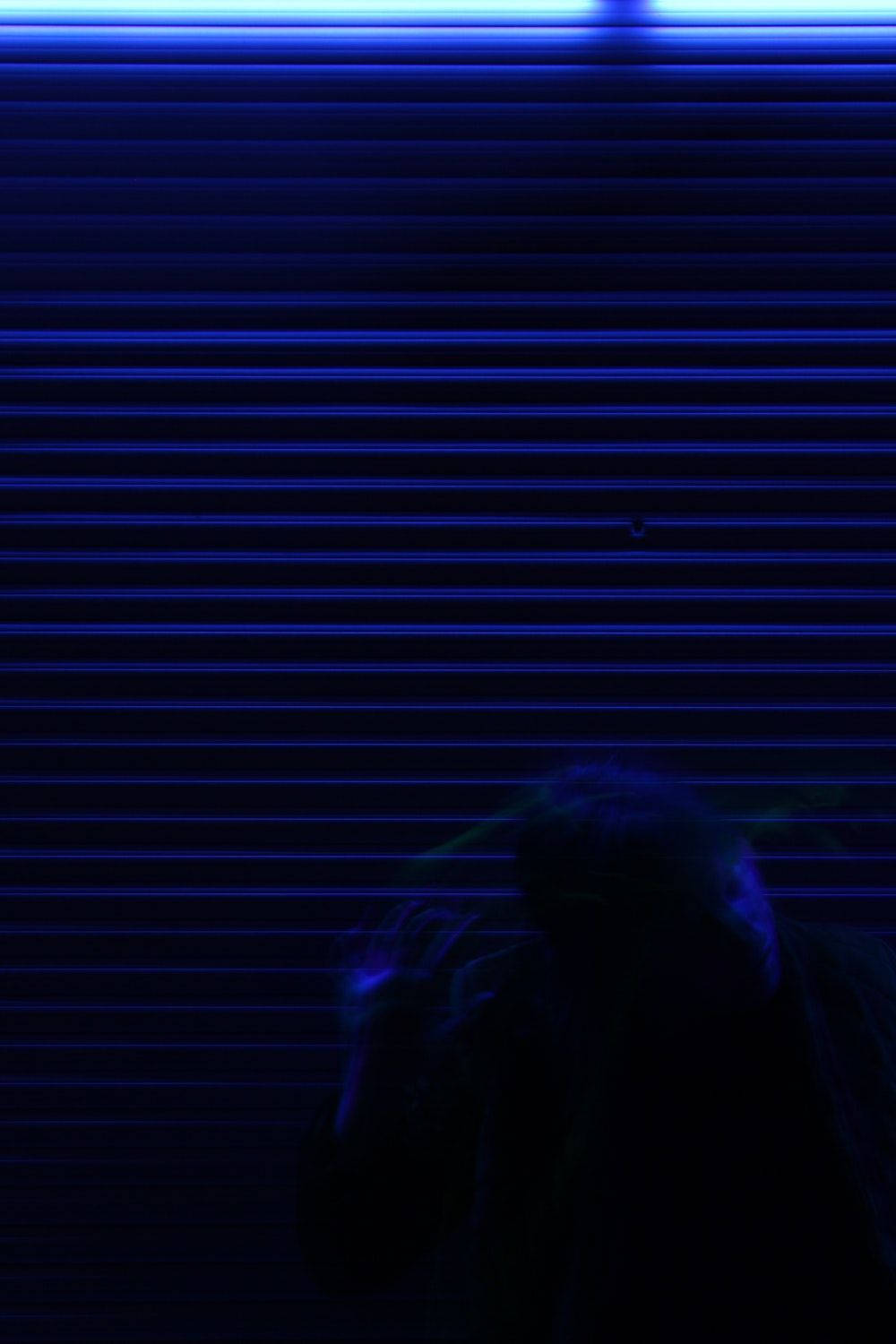 暗黑動漫風格GIF 桌布 壁紙 — 一個陰暗的身影站在昏暗房間中發光的藍色水平百葉窗前。