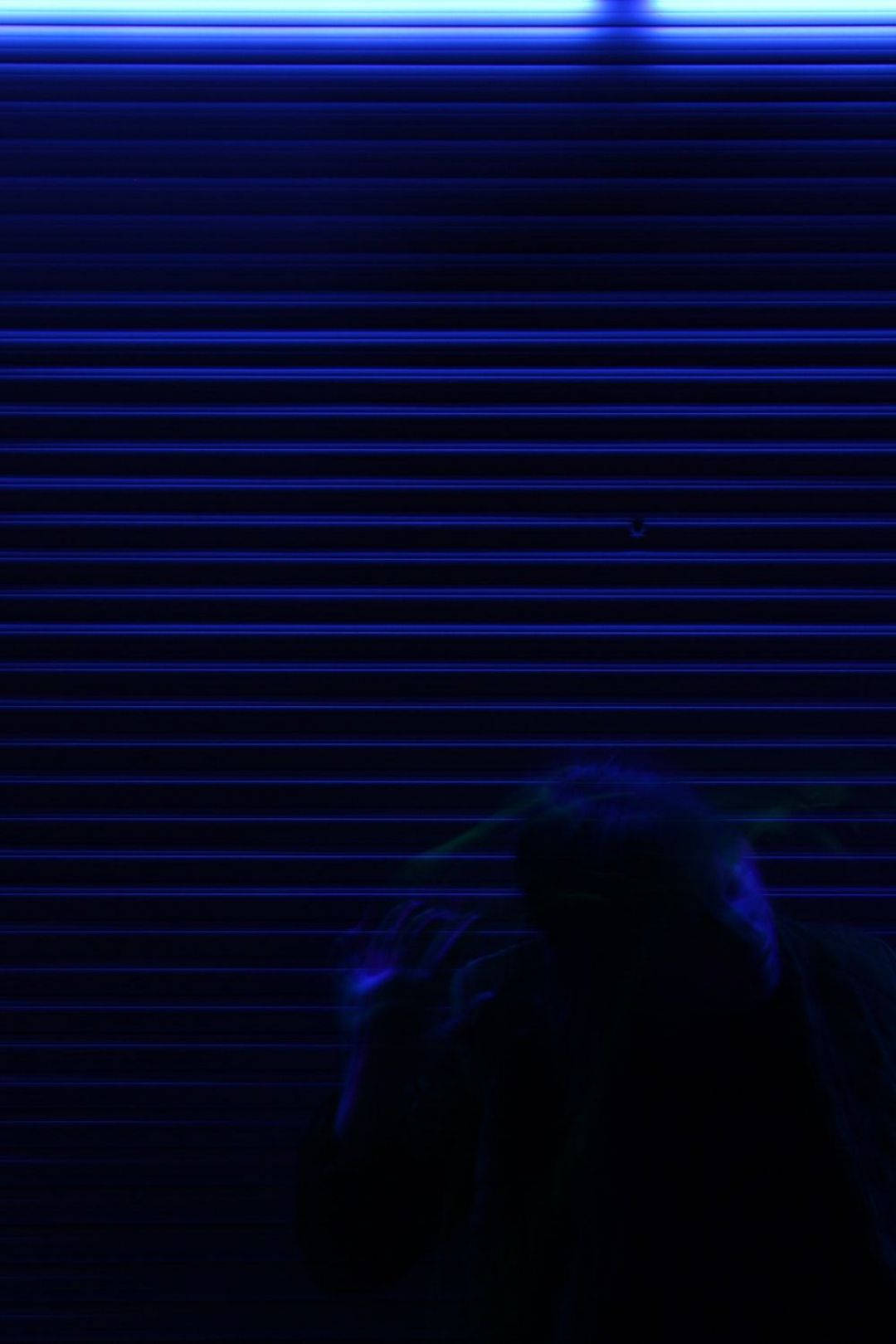 Dark Blue iPhone 桌布 壁紙 — 一個陰暗的身影站在藍色照明的金屬百葉窗前，雙手舉起，處於昏暗發光的城市夜景中。