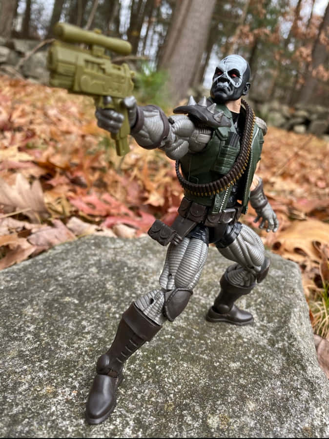 Skullbuster 桌布 壁紙 — 一個陰森的骷髏臉動作人偶，身穿綠色和灰色裝備，在落滿秋葉的森林中，從長滿苔蘚的岩石上瞄準步槍。