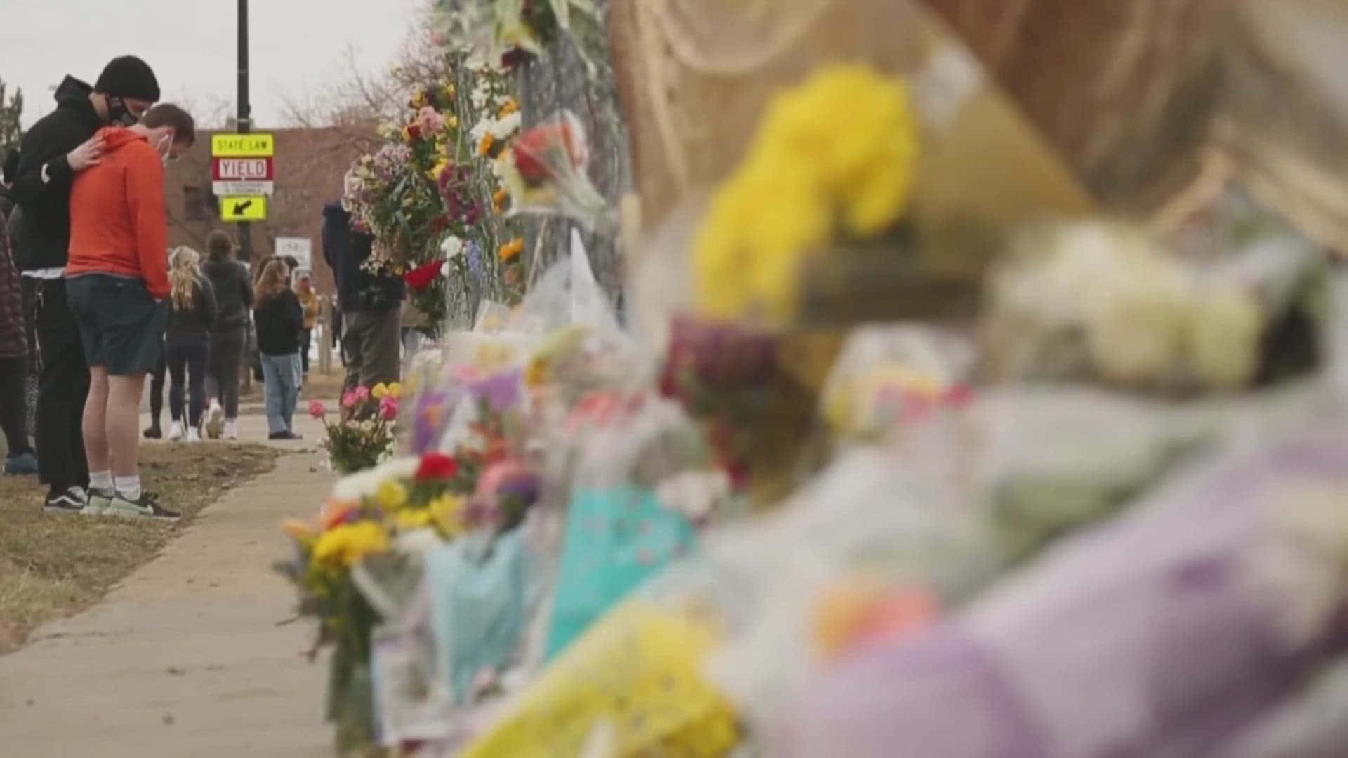 dead 桌布 壁紙 — 一個陰沉的紀念碑，色彩繽紛的花束排列在人行道上，人們靜靜地聚集在一起悼念。