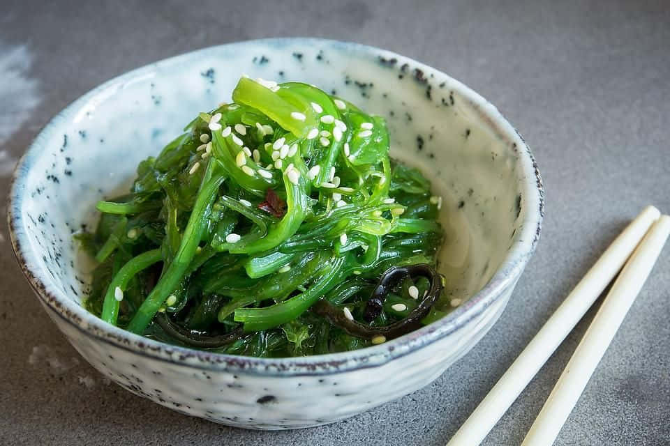 Seaweed 桌布 壁紙 — 一個陶瓷碗裝滿了鮮綠色的海藻沙拉，上面撒上白芝麻，旁邊放著木筷子，放在灰色表面上。