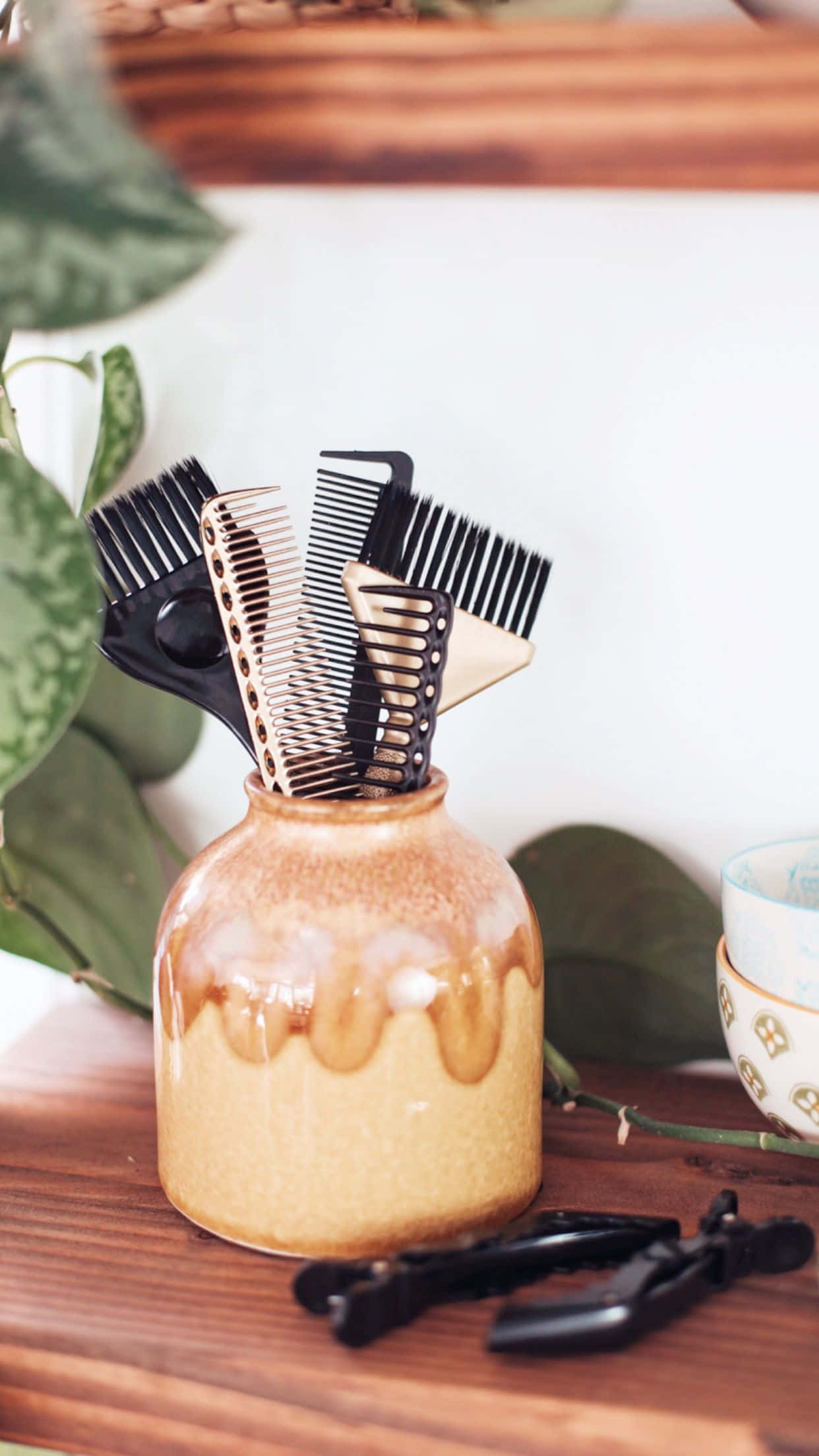 Haircare 桌布 壁紙 — 一個陶瓷花瓶裡放著各種黑色梳子和夾子，放在木質表面上，旁邊有綠葉和一個有圖案的杯子。