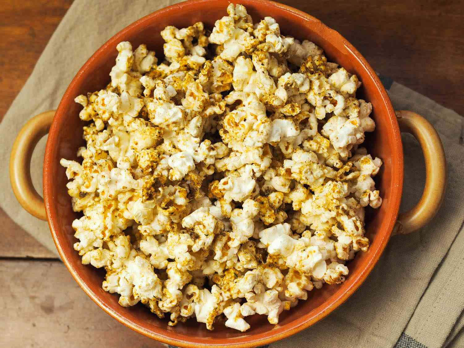 Popcorn 桌布 壁紙 — 一個陶碗盛滿金黃色調味爆米花，放在木質表面的米色布上。