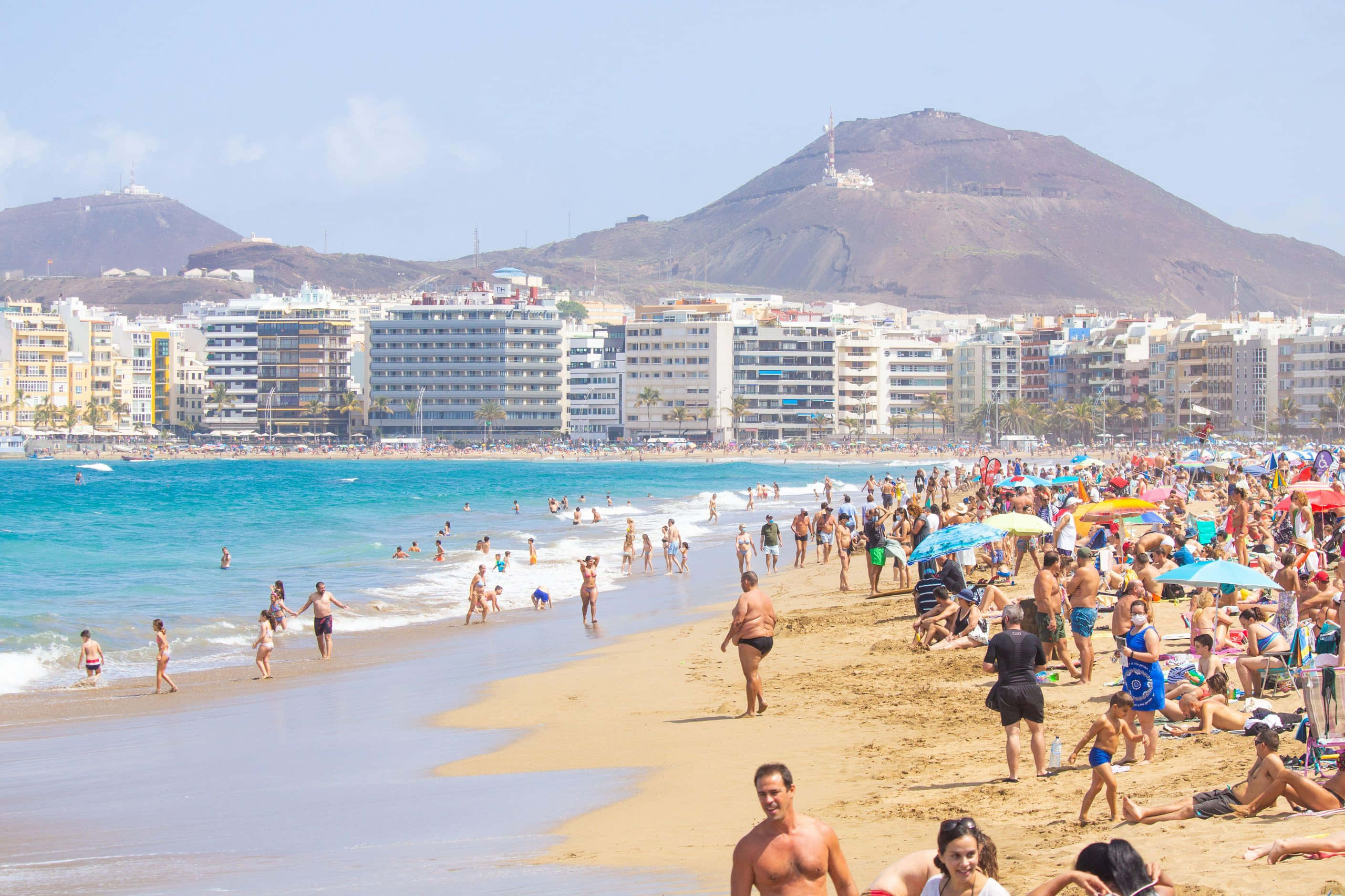 Spanish Beach 桌布 壁紙 — 一個陽光明媚、熙熙攘攘的海灘，擠滿了曬太陽的人和游泳者，背景是清澈的藍天、山脈和酒店。