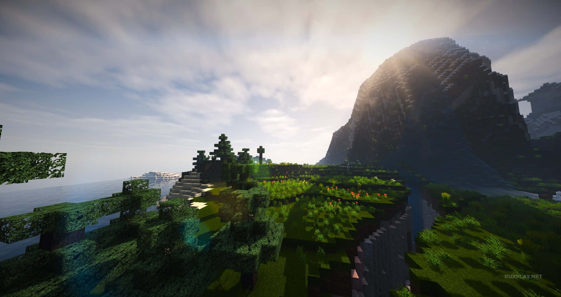 Minecraft Shaders 桌布 壁紙 — 一個陽光明媚的Minecraft景觀，有方塊狀的綠色山丘、樹木，以及一座高聳的山脈，旁邊是平靜的海洋，天空多雲。