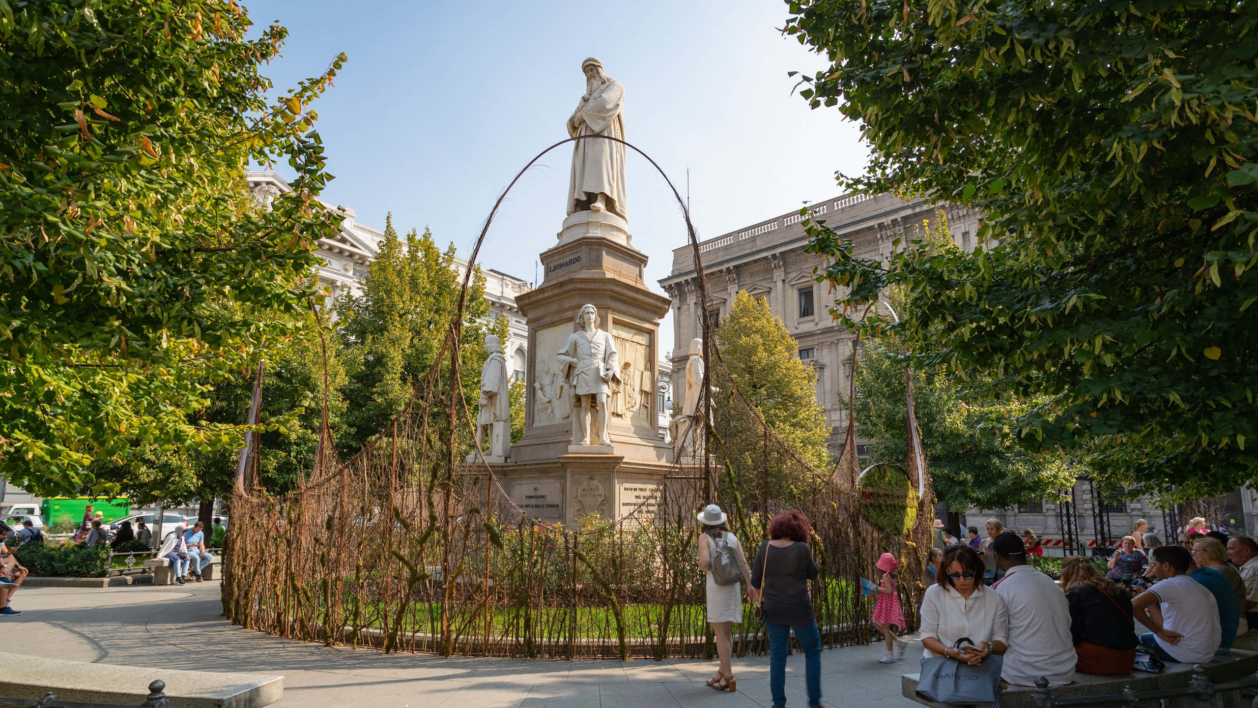 statue 桌布 壁紙 — 一個陽光明媚的城市公園場景，人們在歷史雕像周圍放鬆，四周環繞著茂密的綠樹和古典建築。