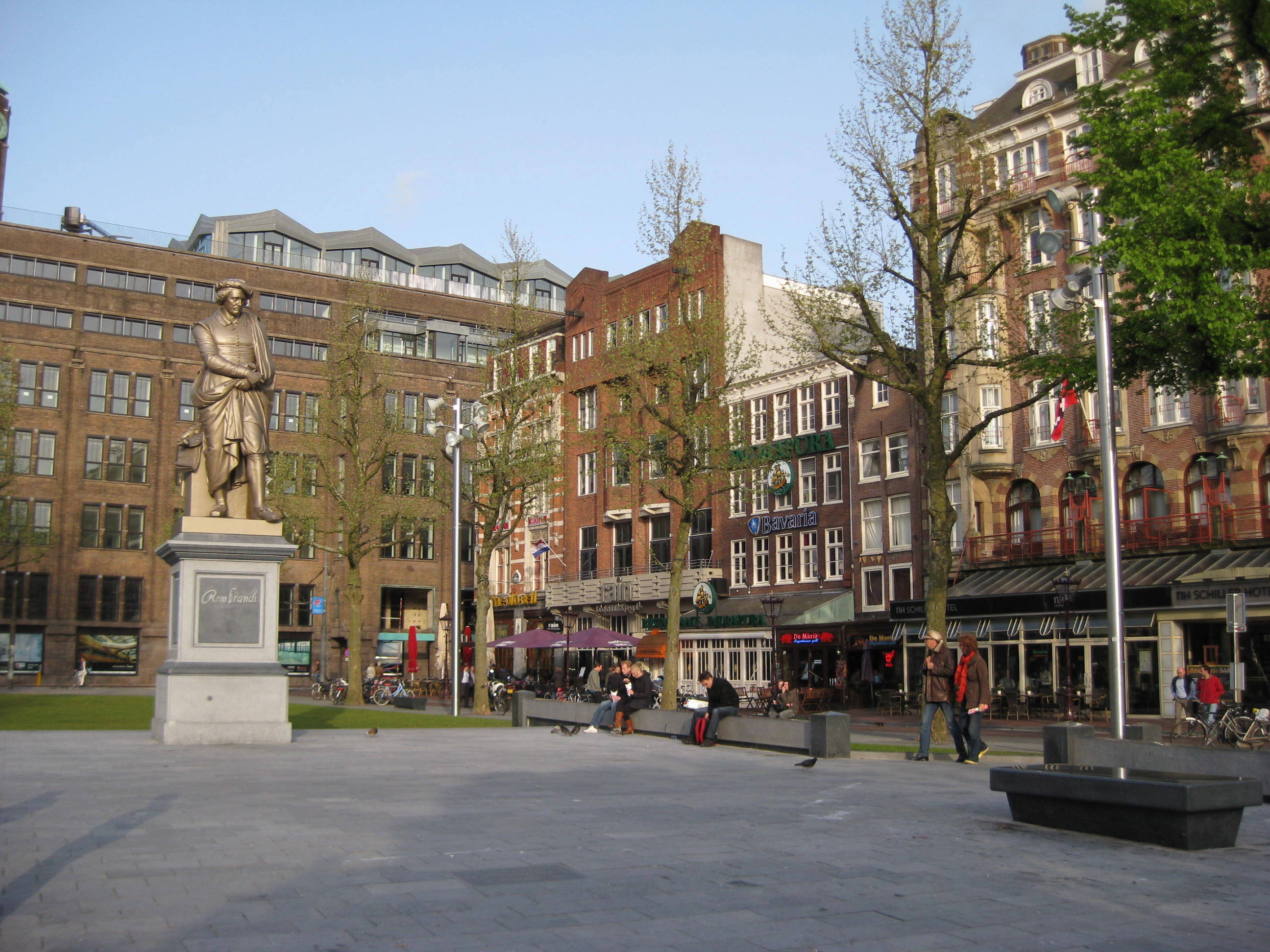 Amsterdam 桌布 壁紙 — 一個陽光明媚的城市廣場，有一座穿著歷史服裝的男性銅像，矗立在磚砌建築前，人們在戶外咖啡館放鬆。